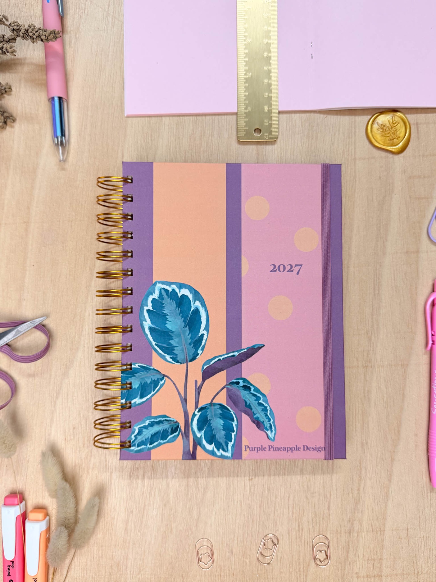 Agenda 2027 com capa colorida, régua, caneta e clips numa mesa de madeira.