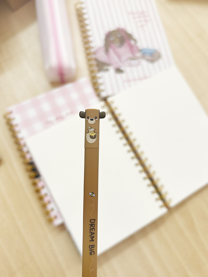 Caneta castanha com ponta em forma de ursinho e cadernos espiralados abertos sobre mesa