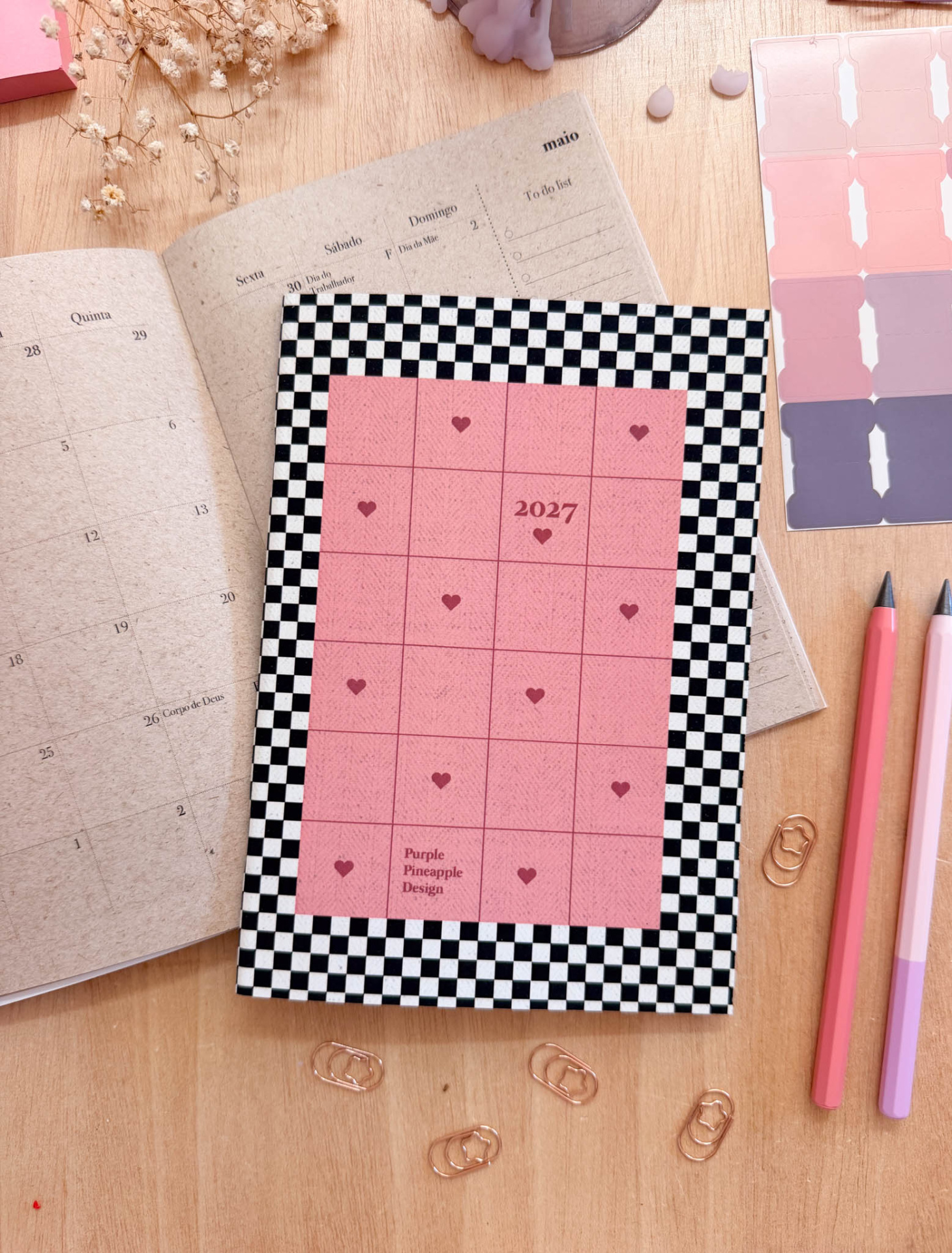 Agenda 2027 com capa xadrez preto e branco e quadrado rosa com corações, sobre mesa de madeira com caderno aberto, clips e lápis.