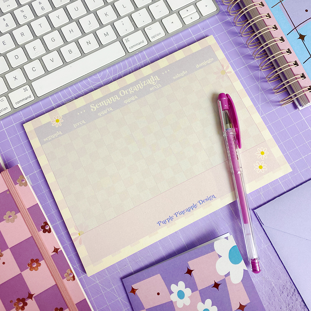 Bloco de notas Semana Organizada com caneta rosa sobre superfície lilás quadriculada e cadernos roxos