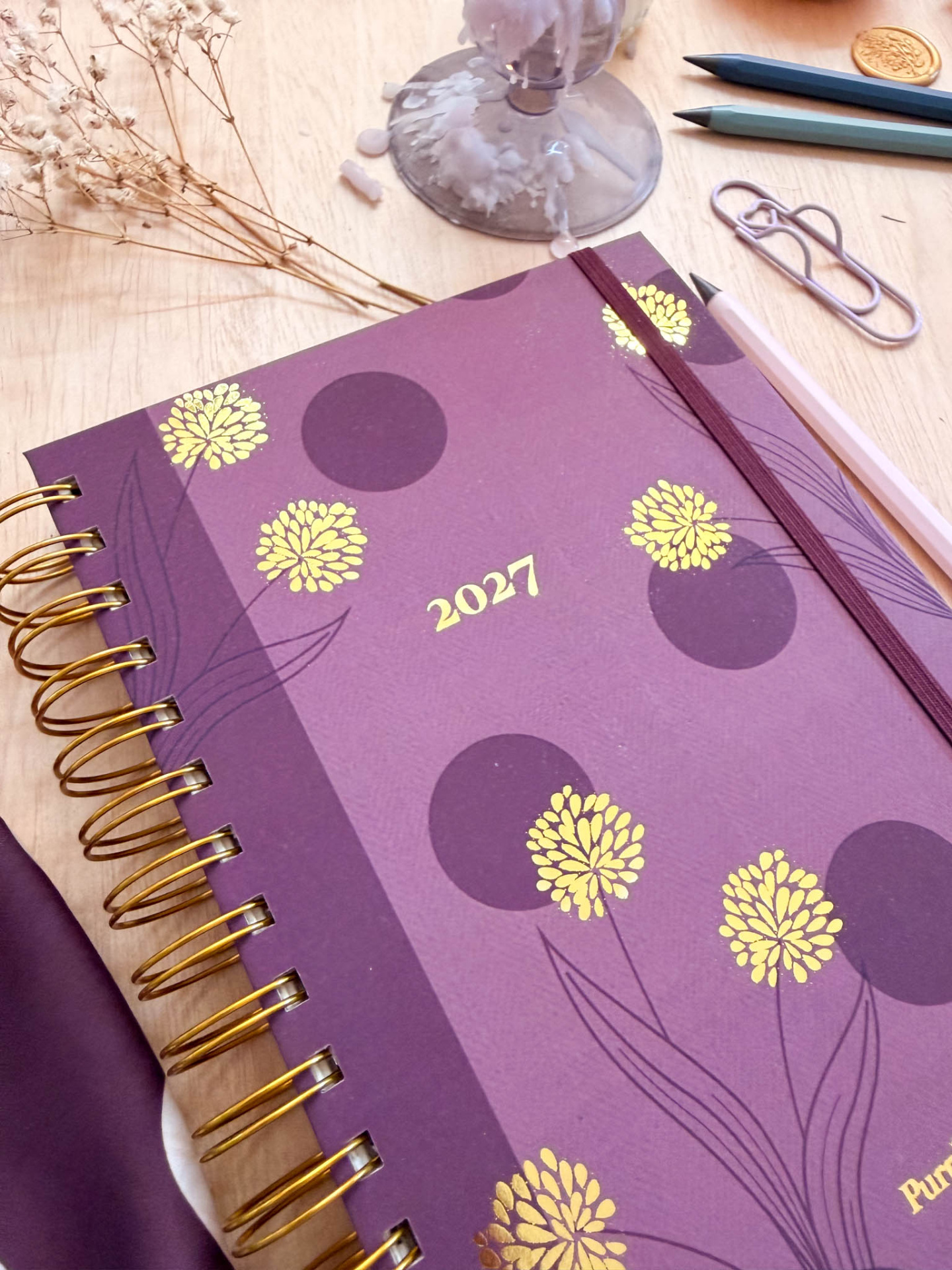 Agenda 2027 roxa com flores amarelas e decoração, rodeada por lápis, clips e flores secas