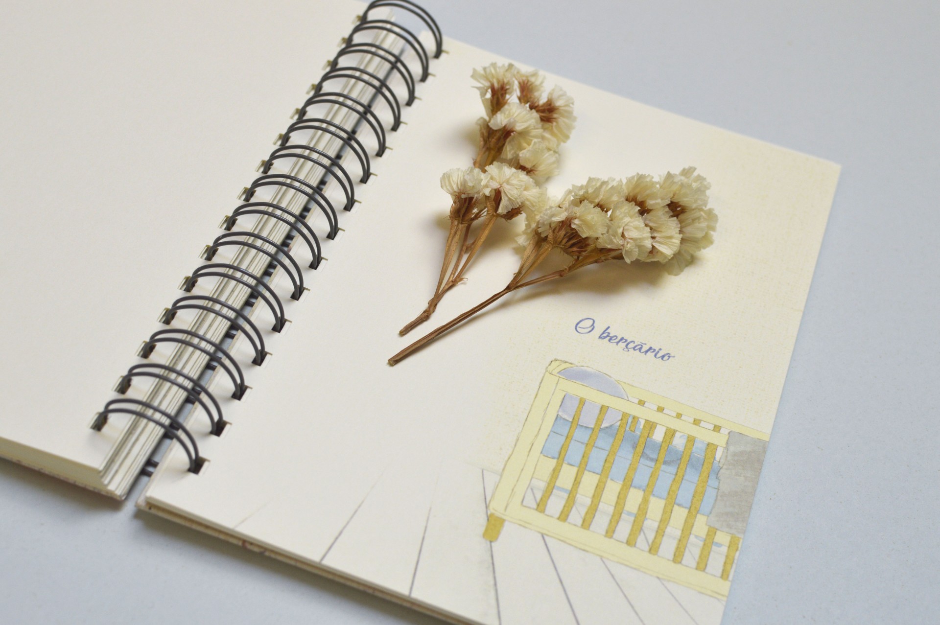 Caderno espiral aberto com ilustração de berço amarelo e flores secas brancas sobre as páginas