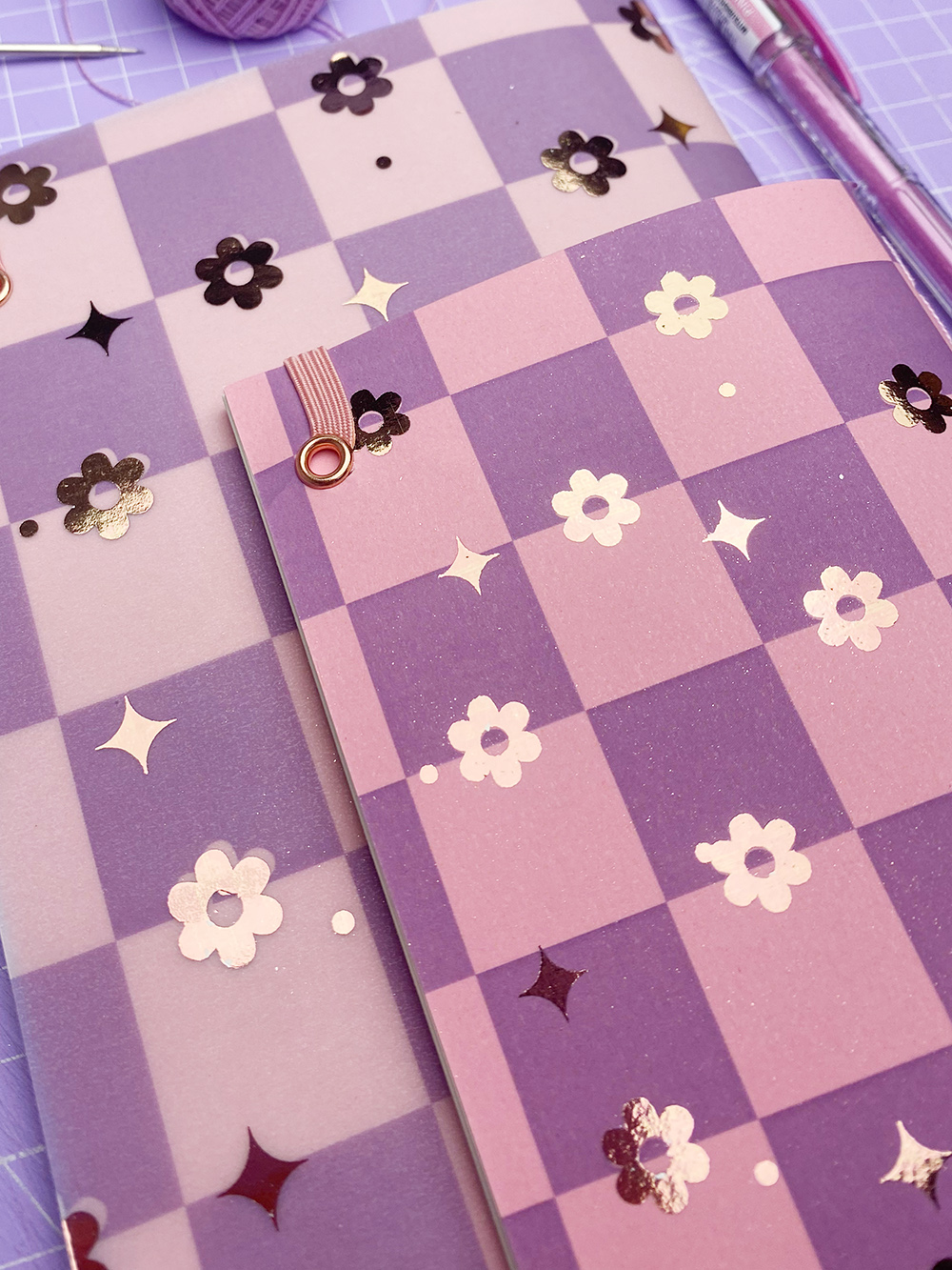 Dois cadernos lilás e rosa com padrões de flores e detalhes metalizados sobre mesa lilás com canetas e linha.