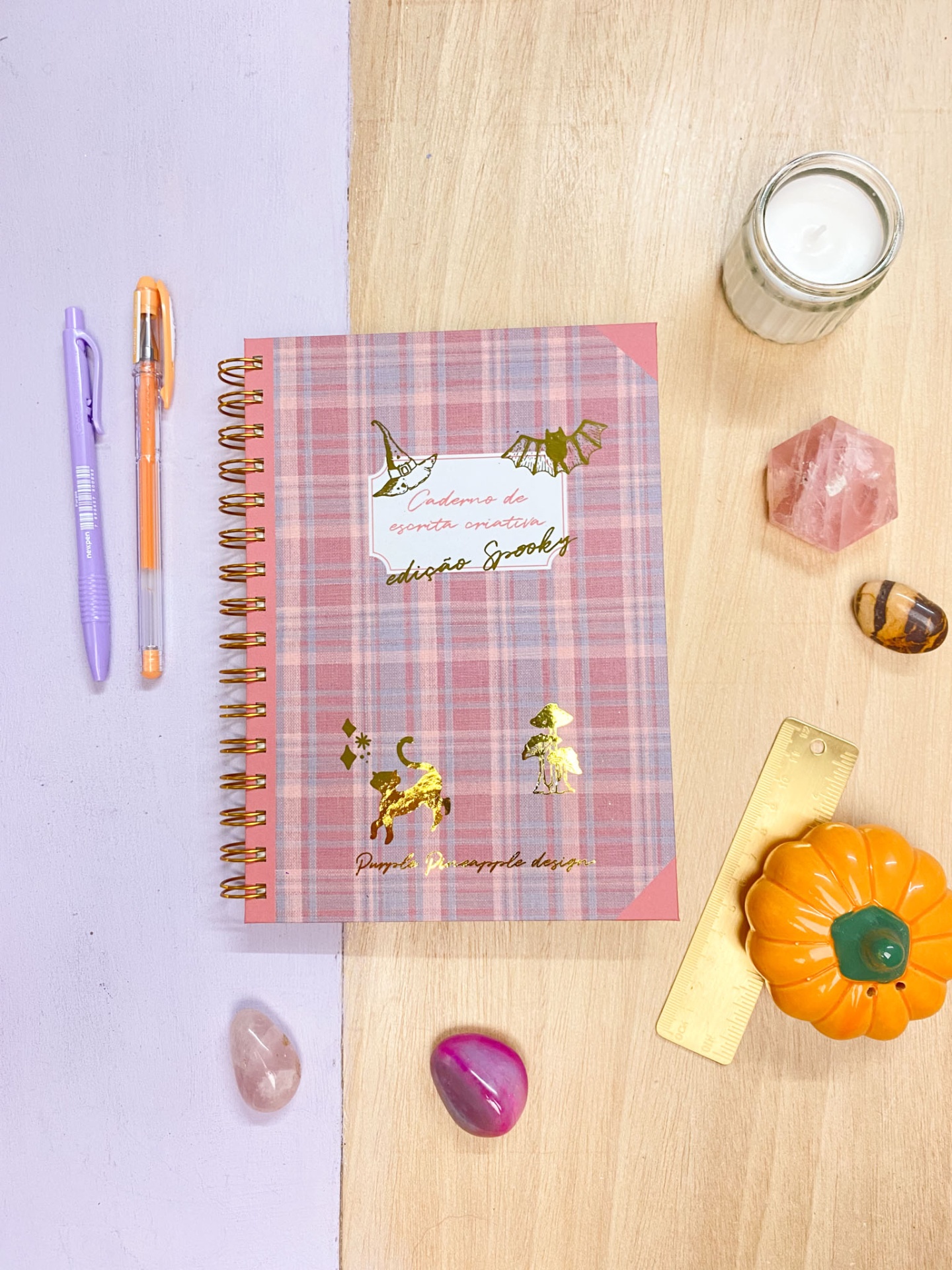Caderno quadriculado rosa com detalhes dourados e canetas, vela, pedras e régua numa mesa