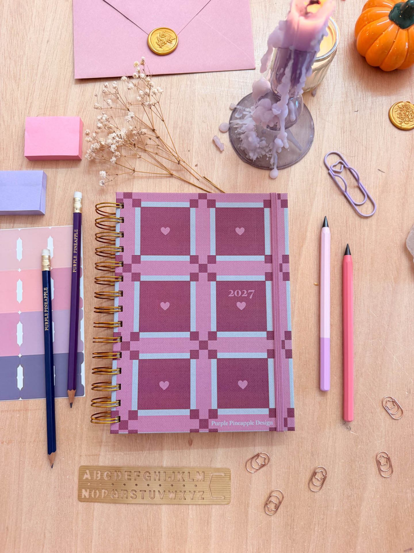 Caderno 2027 rosa e roxo com padrão xadrez e corações, lápis, clips, envelopes selados, régua de madeira e vela derretida.