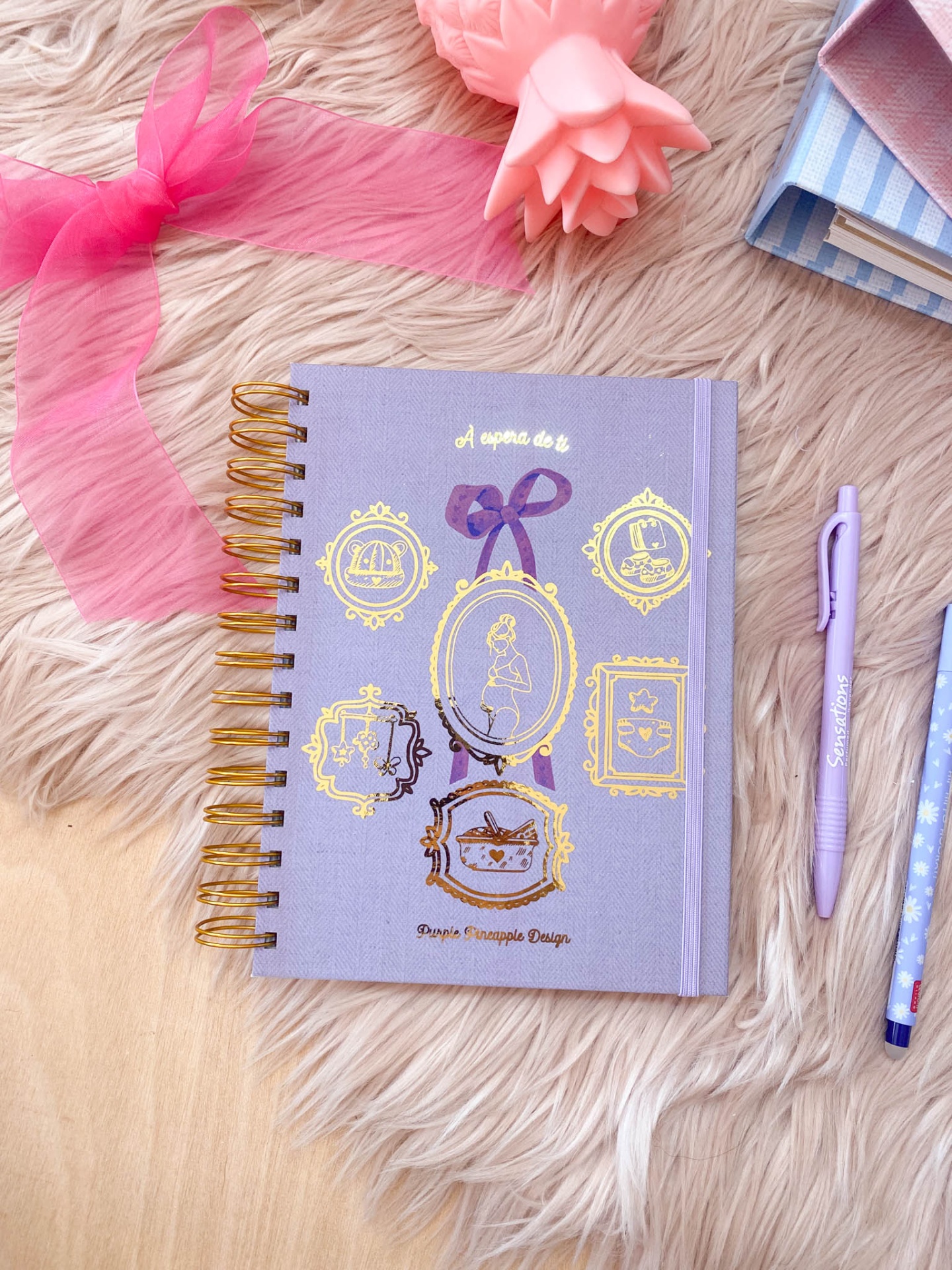 Caderno lilás decorado com molduras douradas, fita rosa e objetos ao redor