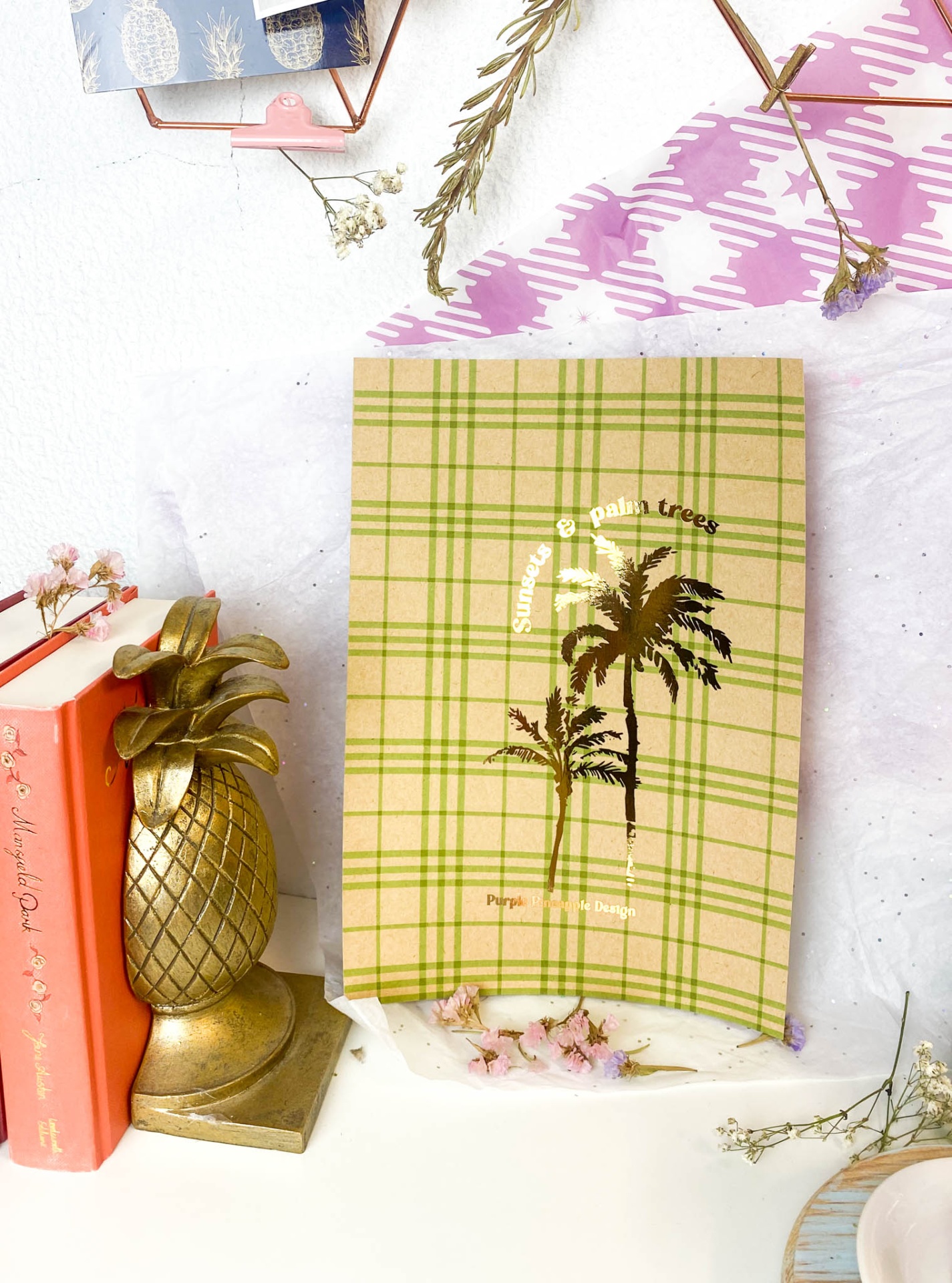 Livro com capa de padrão xadrez verde e bege e texto dourado, ao lado de decoração com abacaxi dourado e livro vermelho