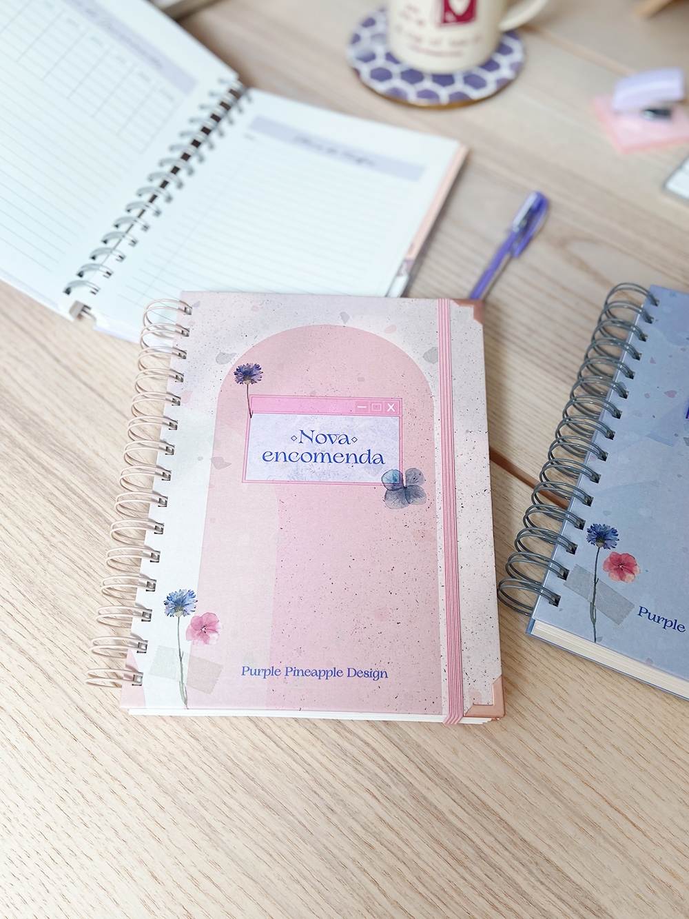 Caderno rosa com flores e texto na capa sobre mesa clara com outros objetos