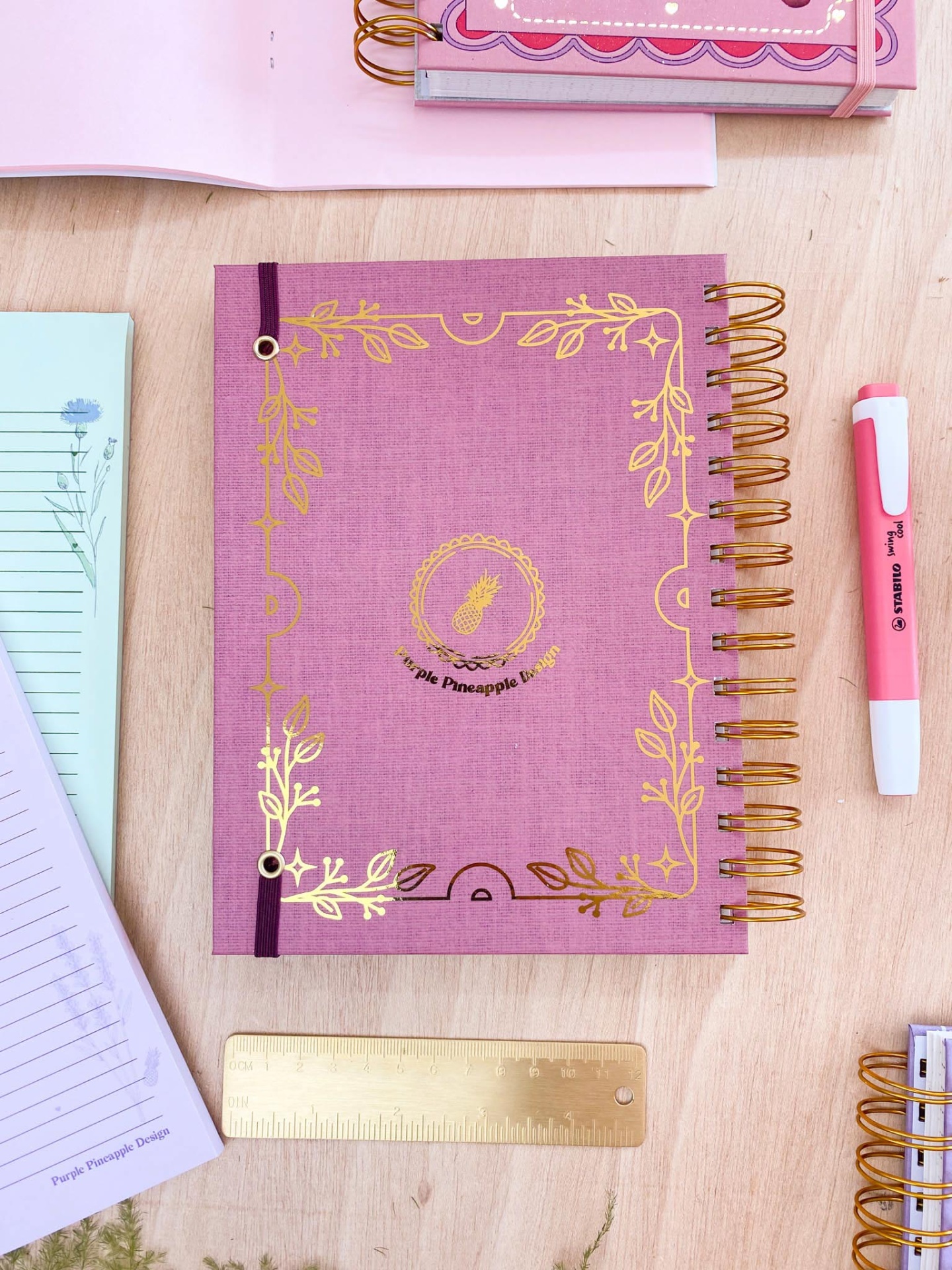 Caderno espiral rosa com detalhes dourados e texto, régua dourada e marcador rosa sobre mesa de madeira