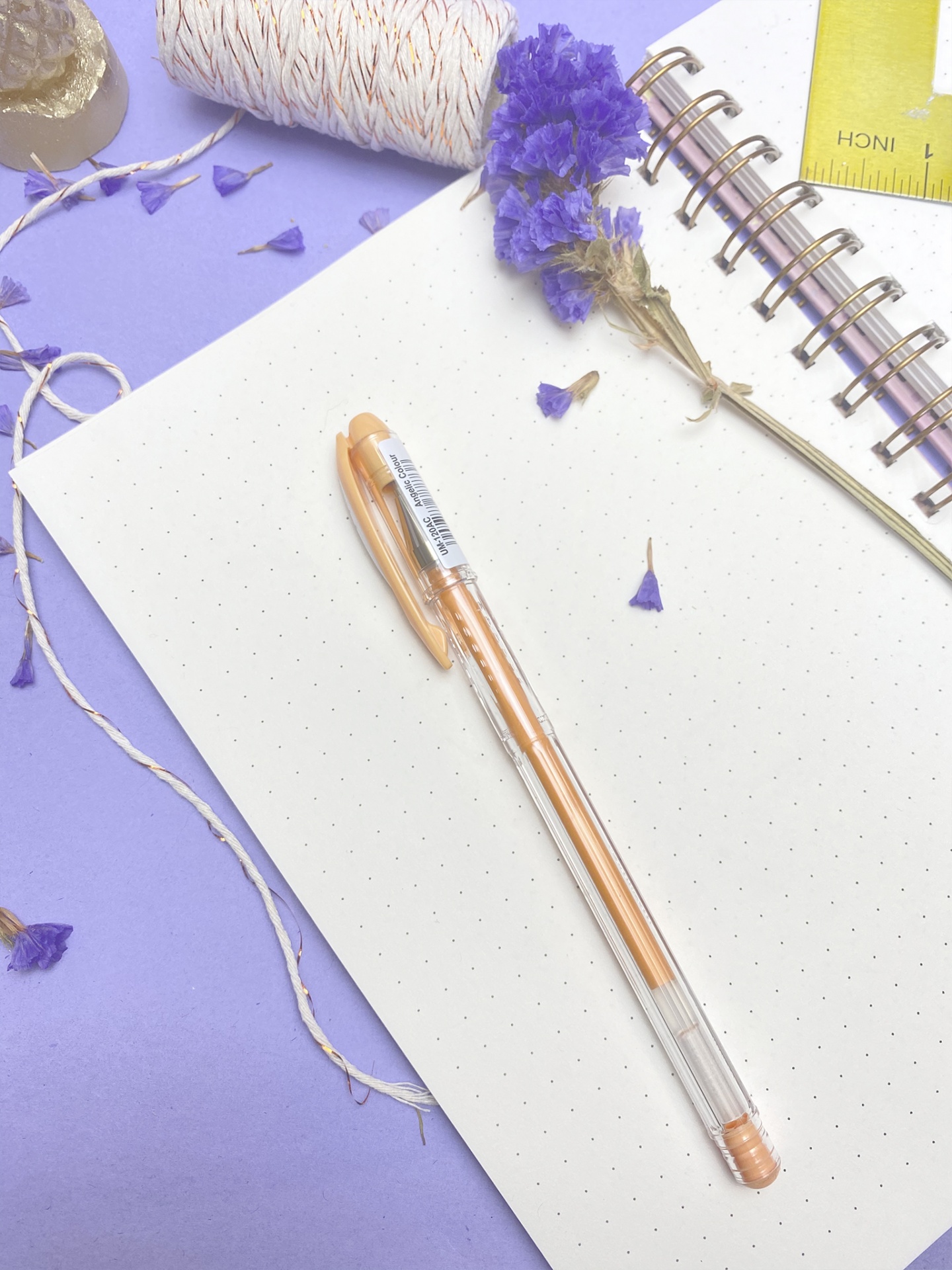 Caderno espiralado com caneta castanha clara, flores secas roxas, cordão e régua amarela sobre superfície lilás