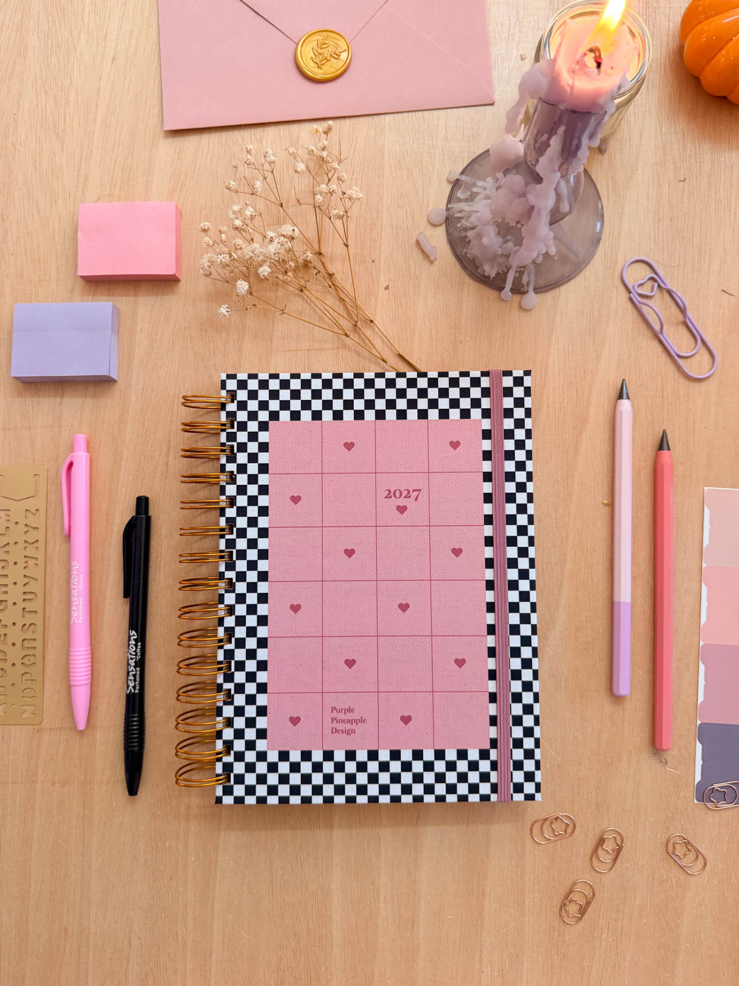 Caderno preto e branco com rosa, canetas, clips e vela rosa derretida num fundo de madeira.