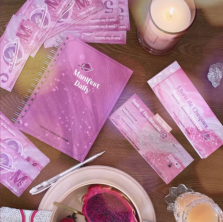 Produtos em tons de rosa incluindo caderno Manifest Daily, notas, vela e dragon fruit sobre mesa de madeira