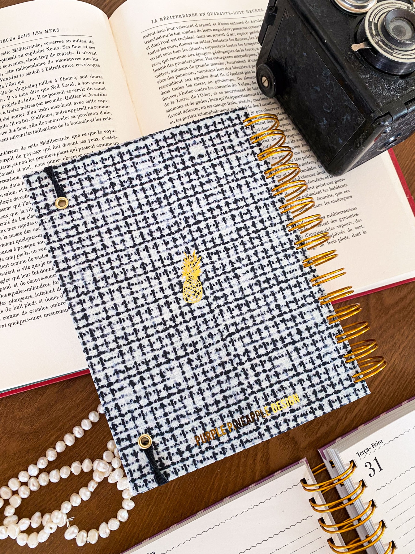 Caderno espiral com capa de tecido quadriculado branco e preto, ananás dourado e texto 'PURPLE PINEAPPLE DESIGN' sobre livro aberto e máquina de escrever.
