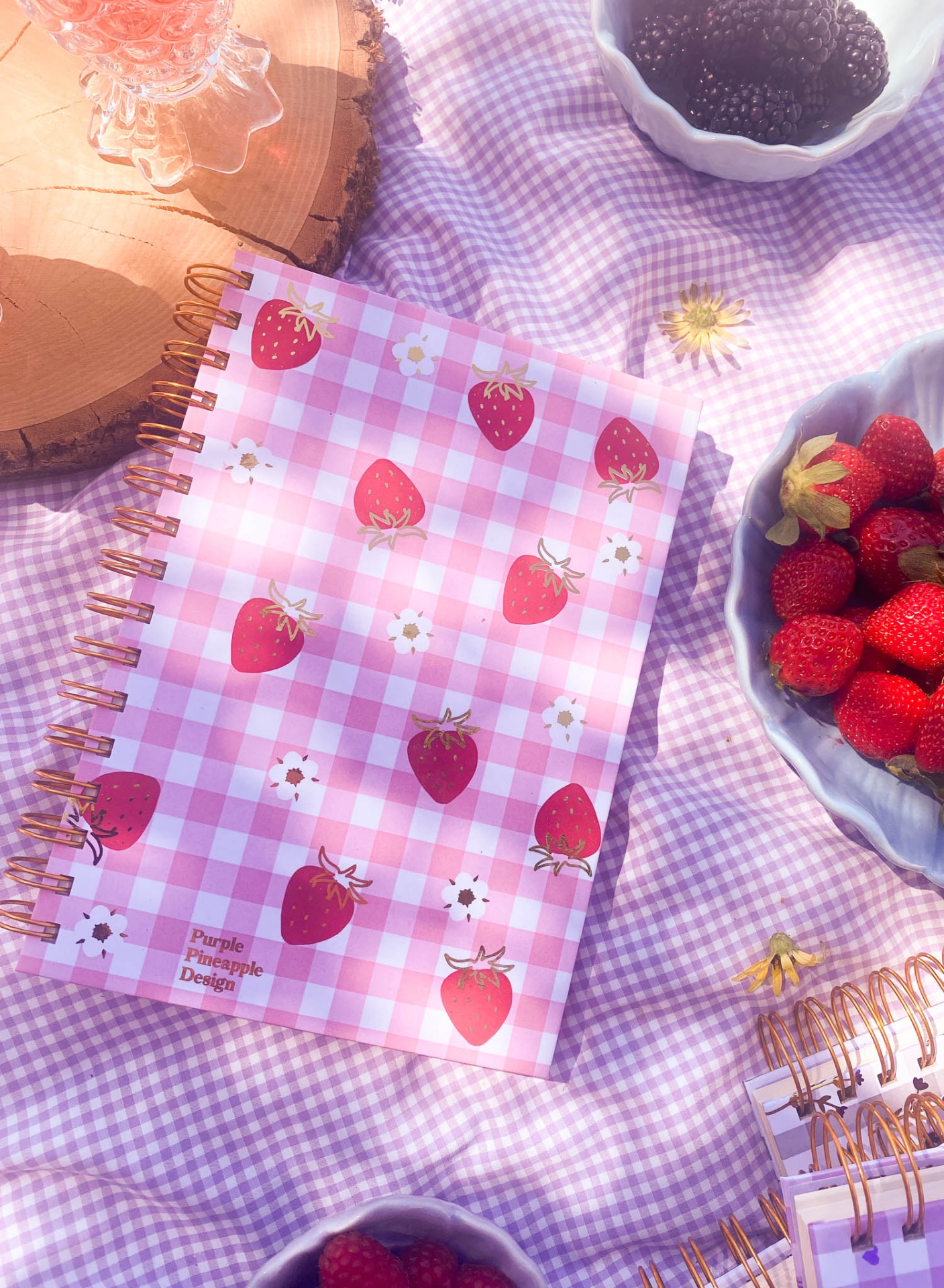 Caderno rosa xadrez com morangos sobre toalha lilás xadrez e frutas em tigelas