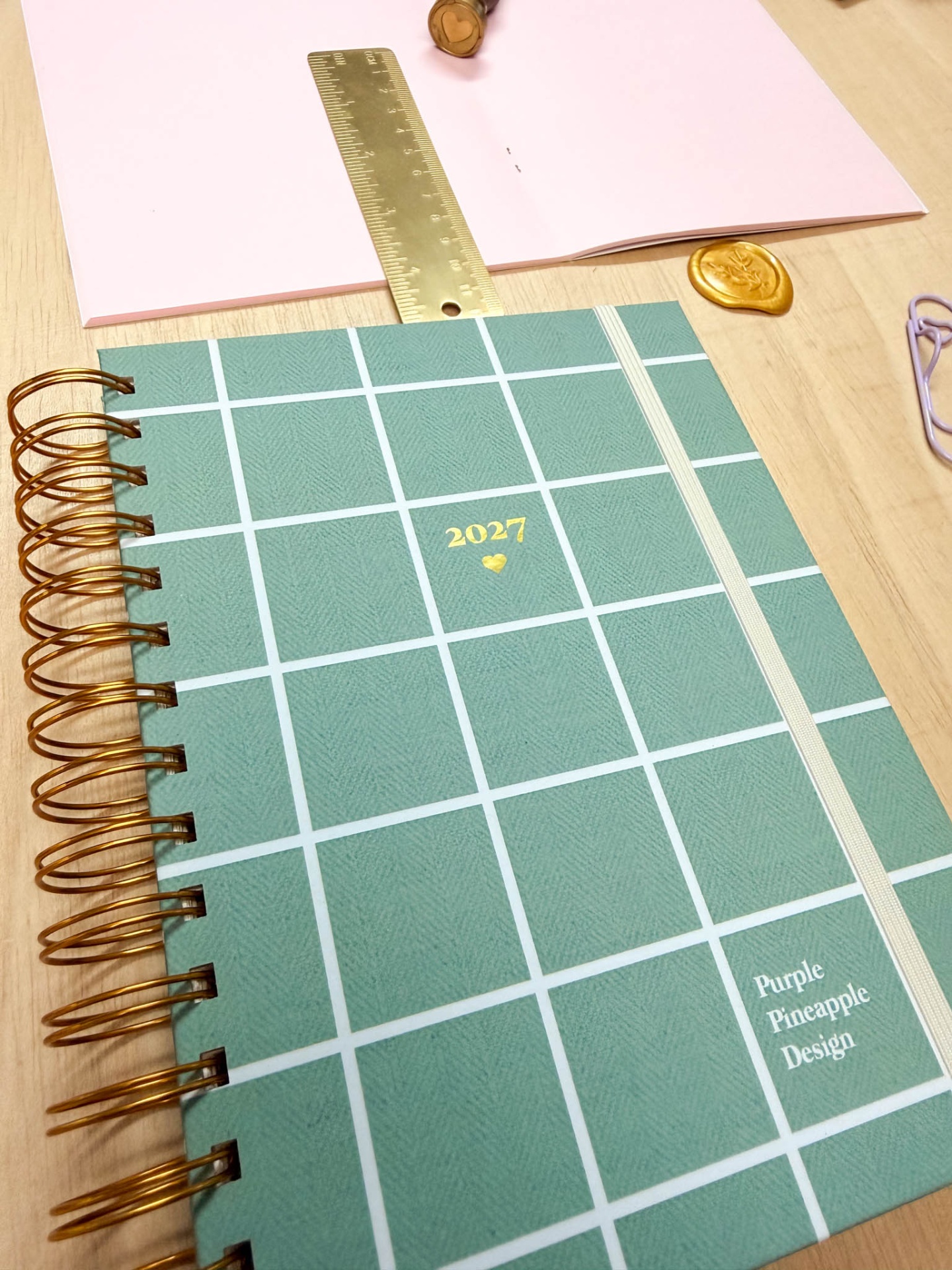 Agenda espiral verde com elástico e texto 2027 e Purple Pineapple Design sobre mesa de madeira