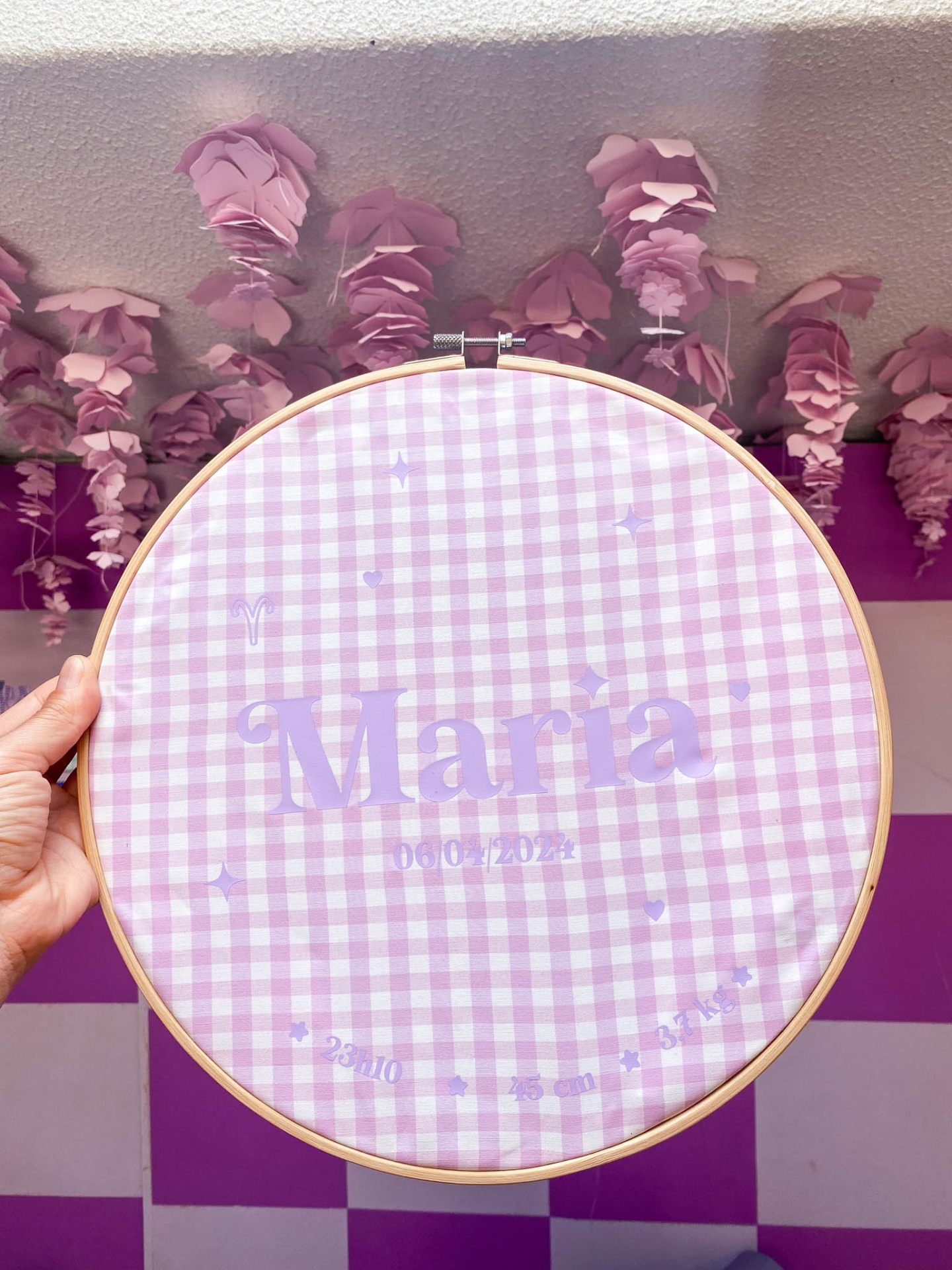 Bastidor de tecido xadrez lilás e branco com texto Maria e data, com flores rosa ao fundo