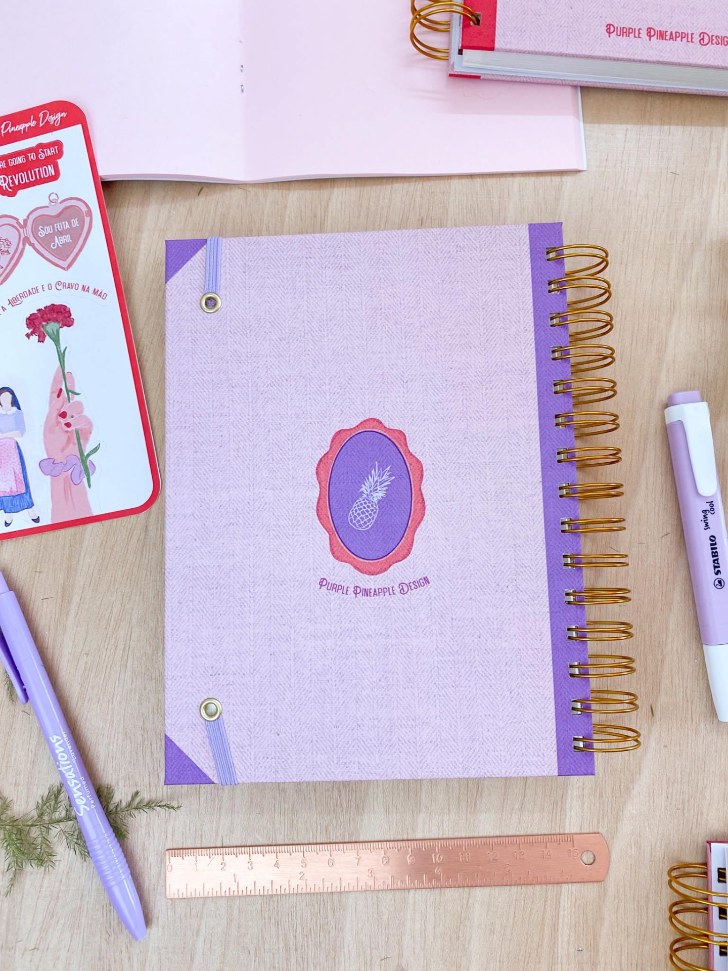 Caderno com capa rosa claro e detalhes roxos, selo oval com ananás, régua rosa e canetas lilás sobre mesa de madeira