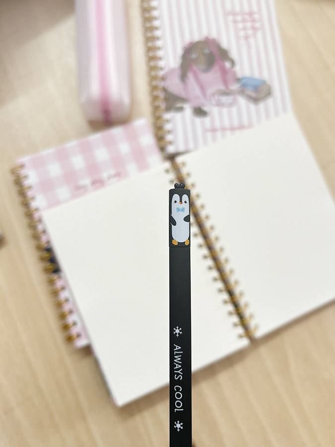 Lápis preto com pinguim e texto Always Cool sobre mesa com cadernos e estojo