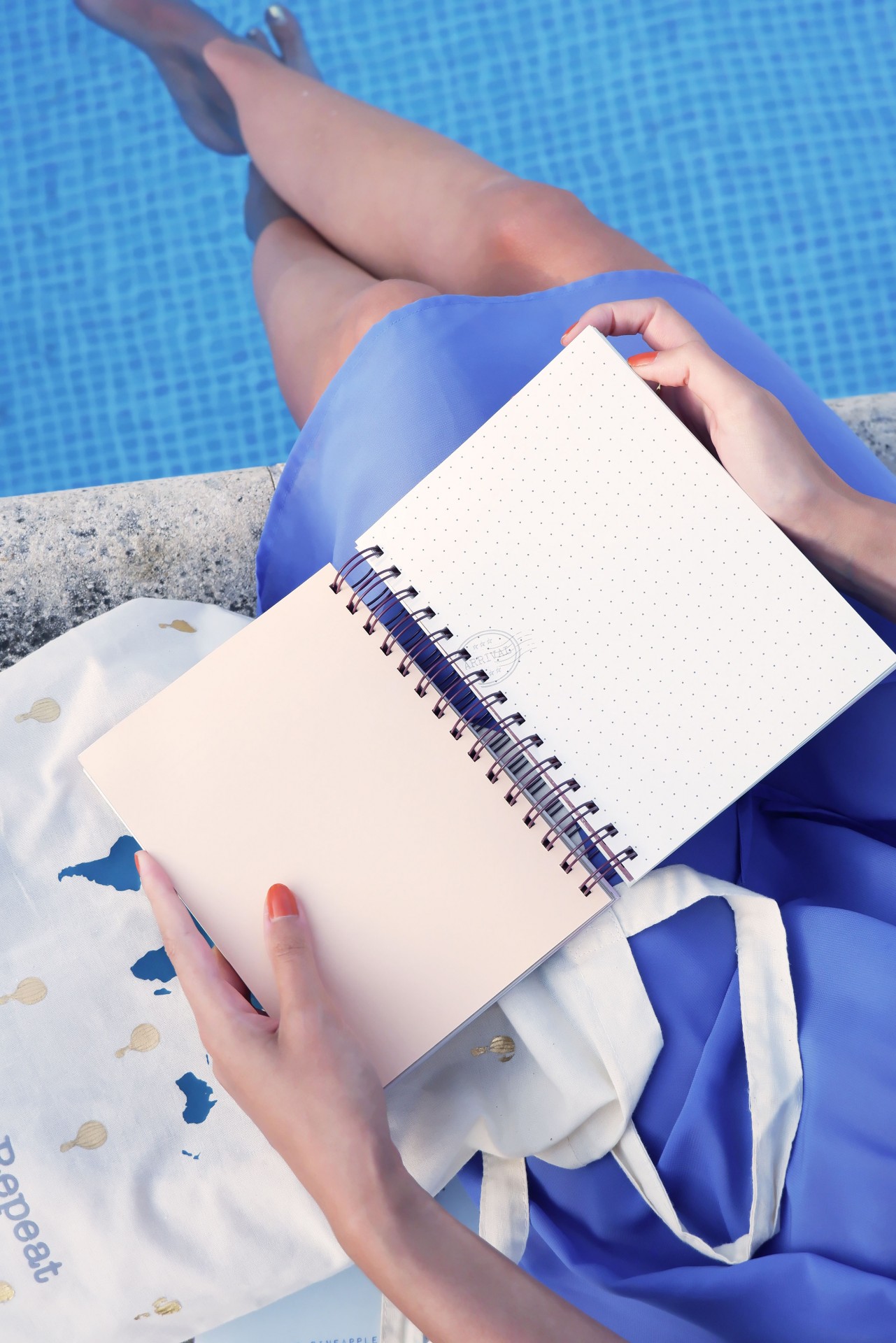 Pessoa segurando caderno de espiral com páginas em ponto junto a piscina, com sacola de tecido ao lado