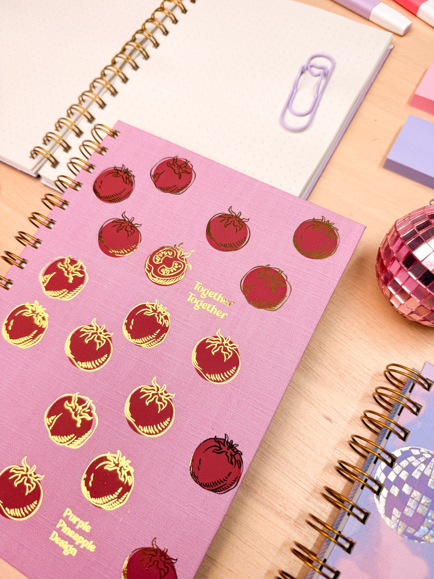 Caderno rosa com desenhos de tomates dourados e vermelhos numa mesa com outros cadernos e acessórios
