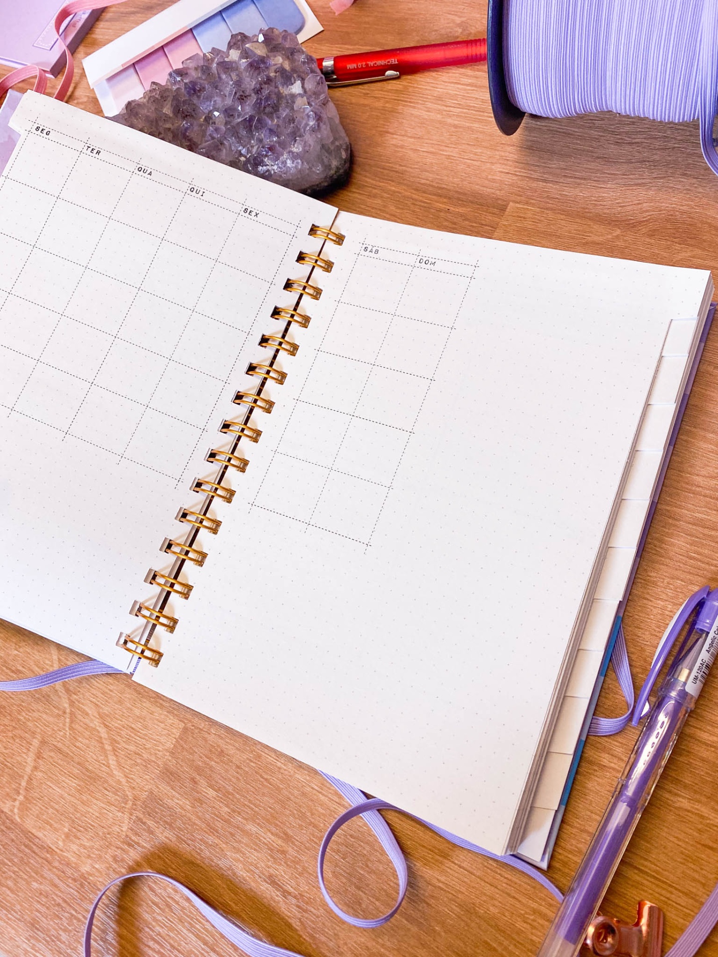 Caderno aberto de capa lilás com espiral dourada sobre mesa de madeira com cristal, canetas e fita