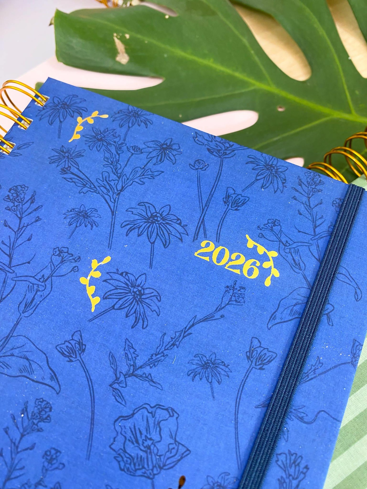 Agenda azul com flores e detalhes amarelos, elástico e espiral dourados sobre folha verde