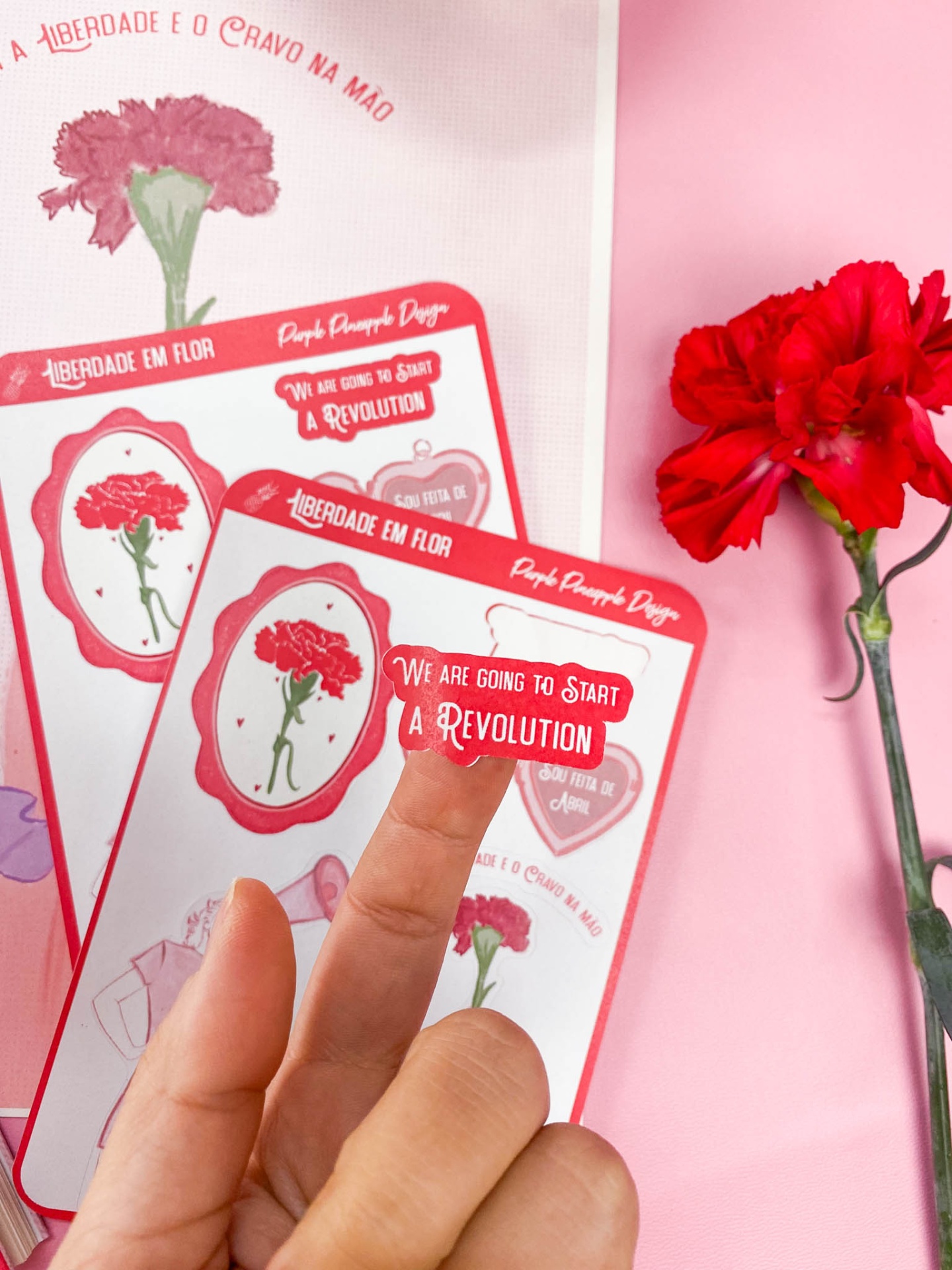 Adesivos com cravo vermelho e frases sobre fundo rosa com uma mão segurando um adesivo e flor real vermelha ao lado