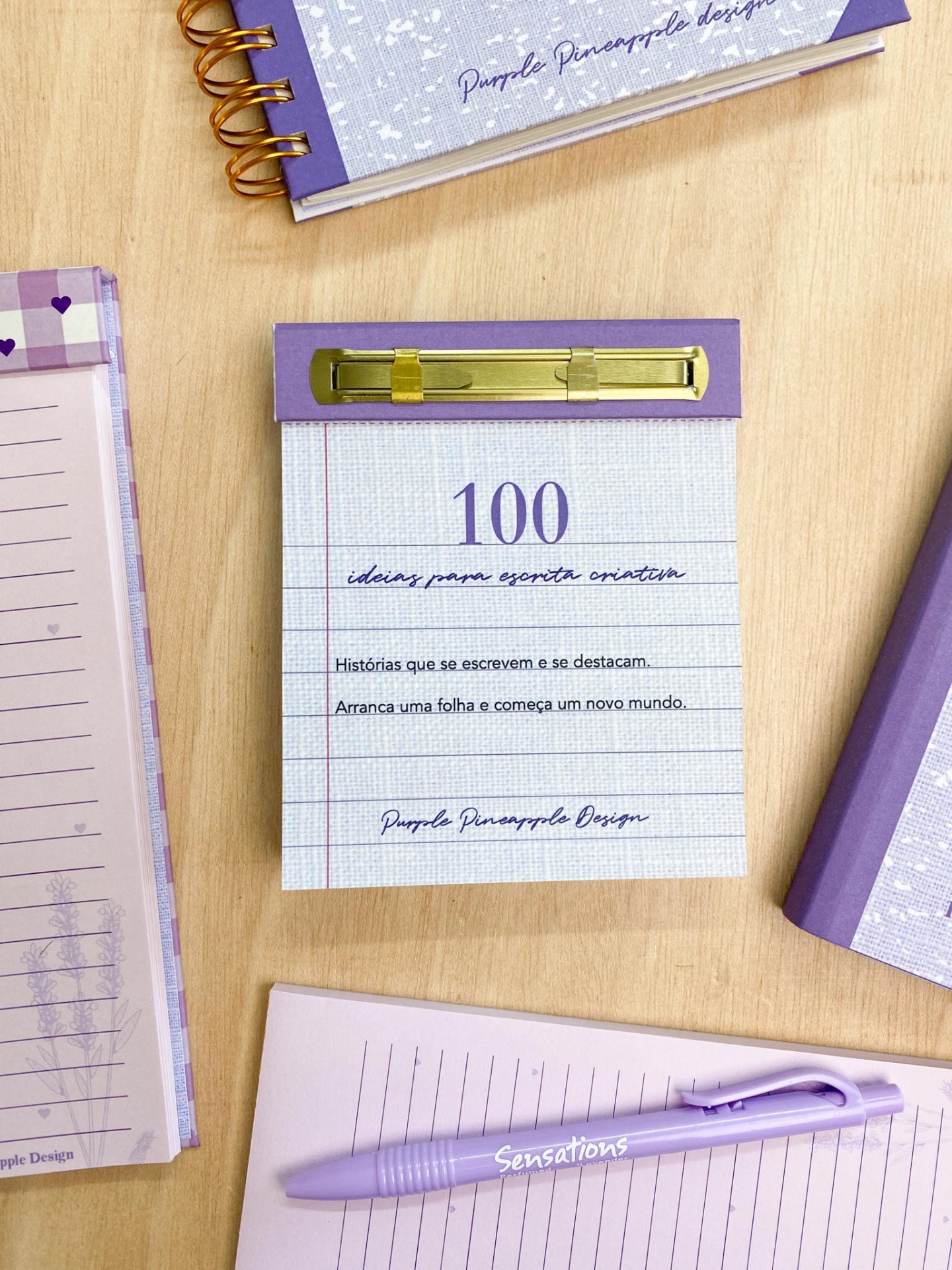 Bloco de notas roxo com prendedor dourado e texto em português, rodeado por materiais de escrita lilás numa mesa de madeira.