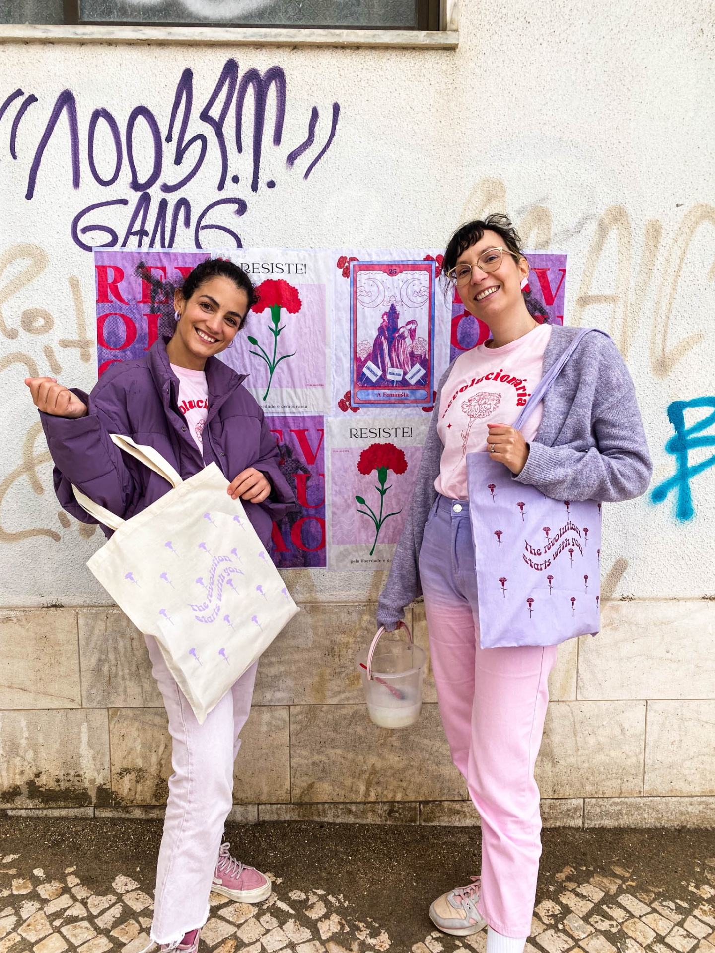Duas mulheres segurando sacos de pano estampados em frente a muro com grafitis e cartazes