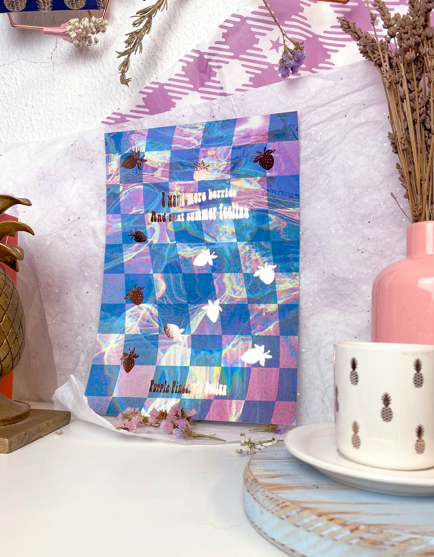 Cartaz azul e roxo quadriculado com insetos, caneca branca com abacaxis, vaso rosa e flores secas em fundo claro