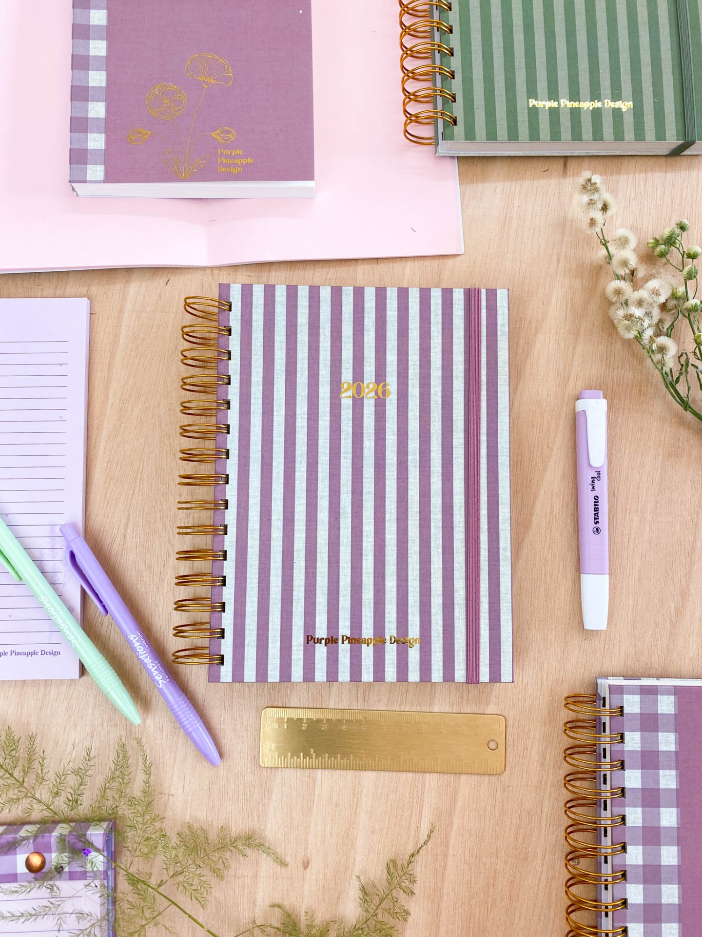 Conjunto de cadernos e agendas espiraladas listradas em roxo, lilás e verde com acessórios e flores sobre mesa de madeira clara