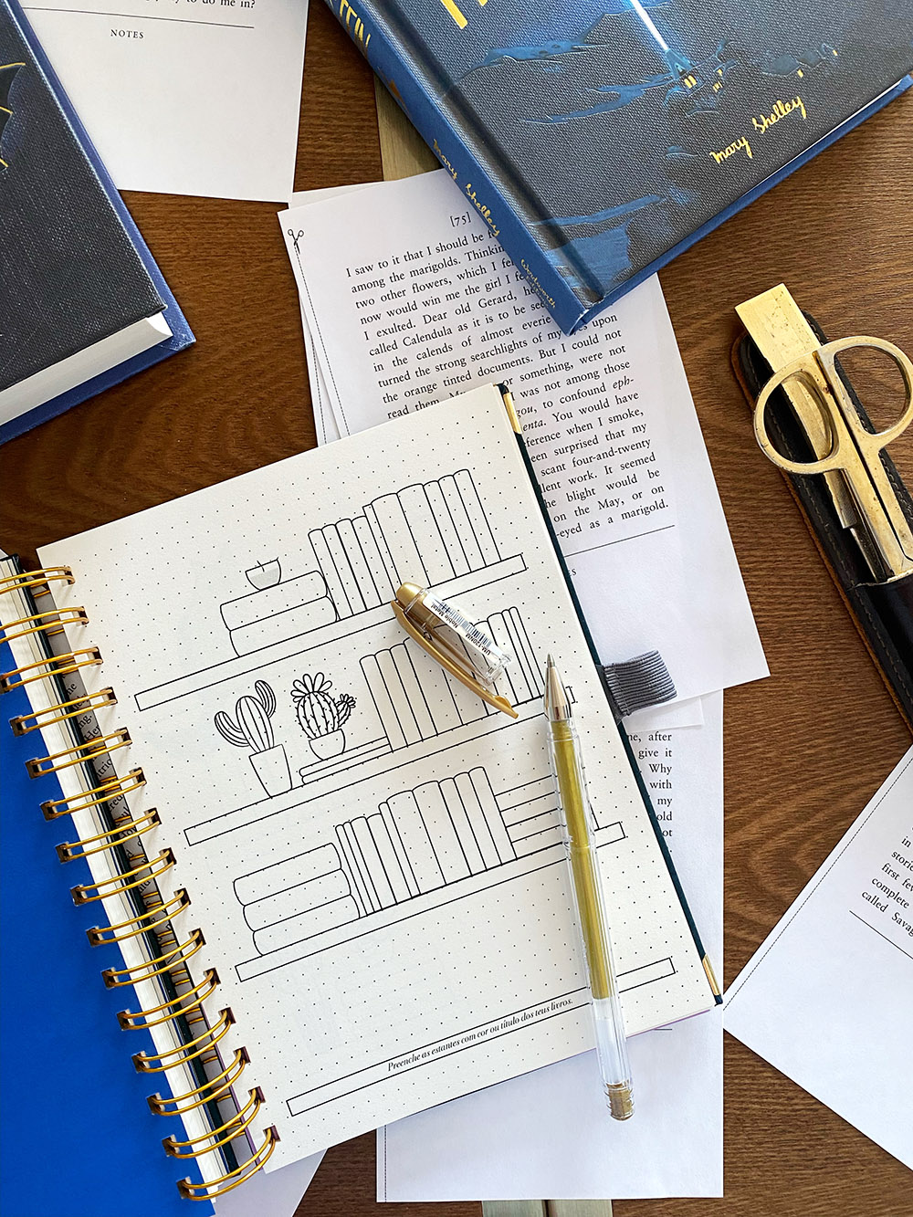 Mesa com caderno azul espiral, papéis, caneta e estojo com tesoura dourada