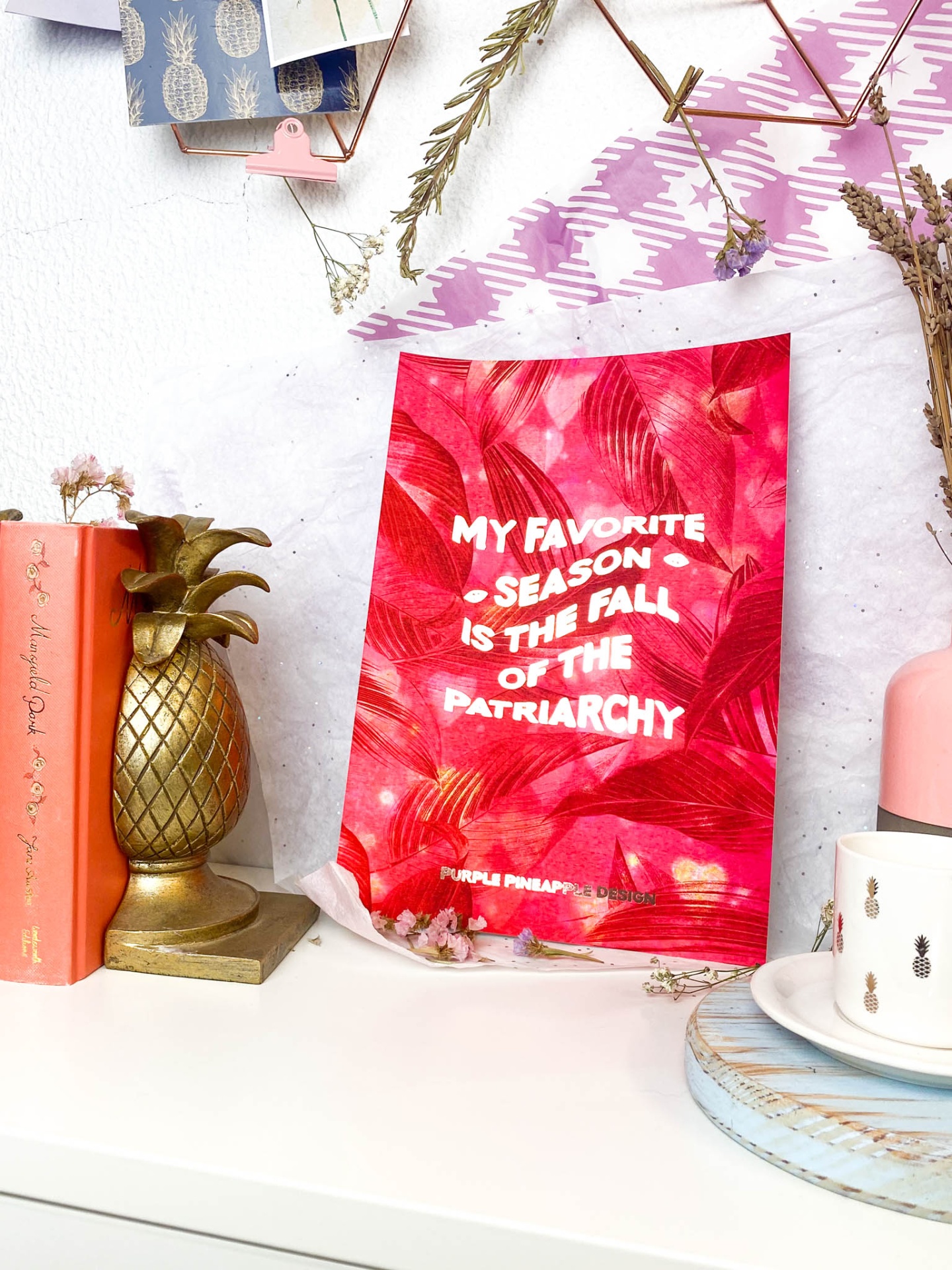 Poster vermelho com frase branca na mesa decorada com livro, suporte abacaxi, flores secas e jarro rosa