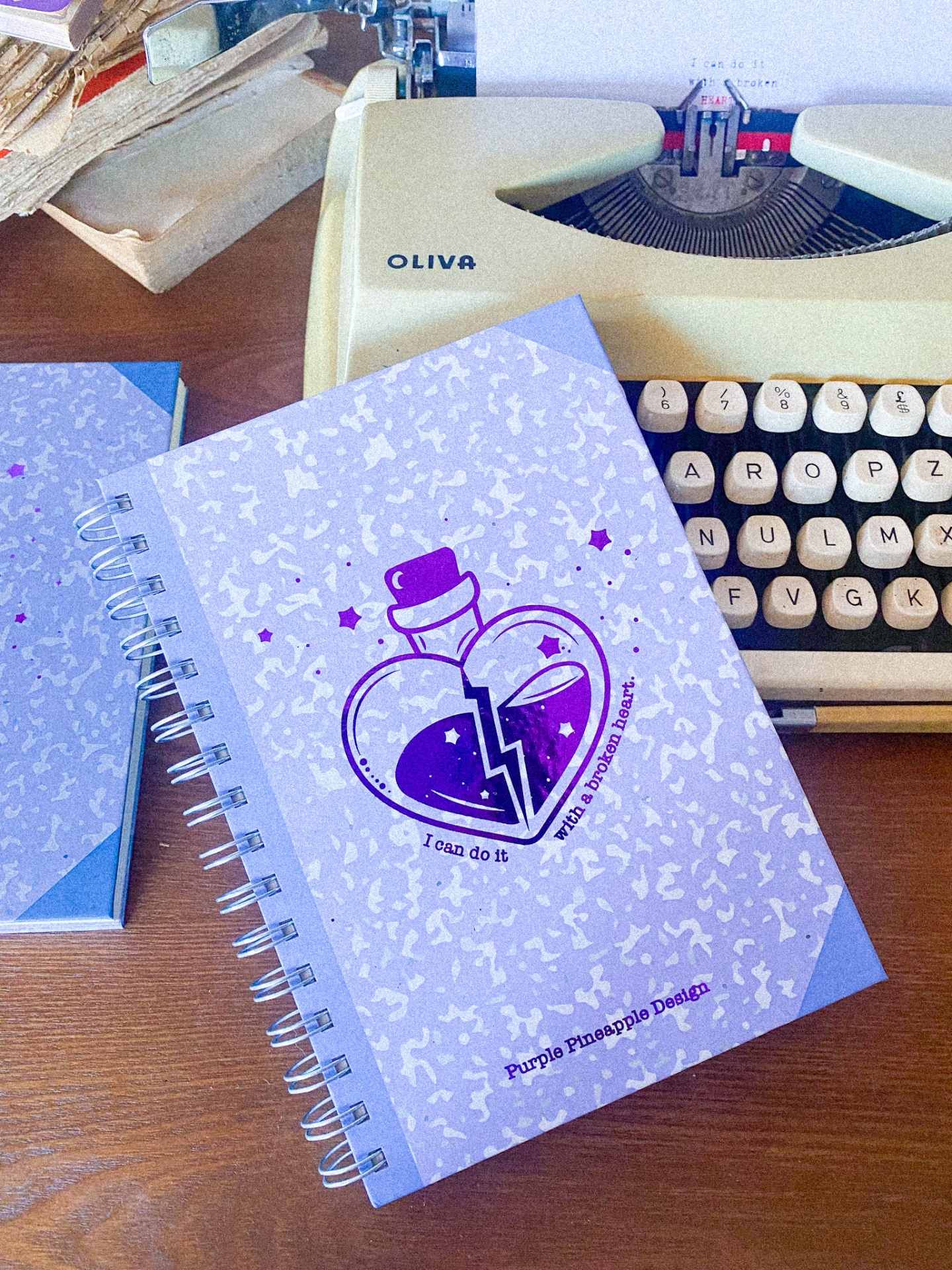 Caderno roxo espiralado com ilustração de coração partido e máquina de escrever cream