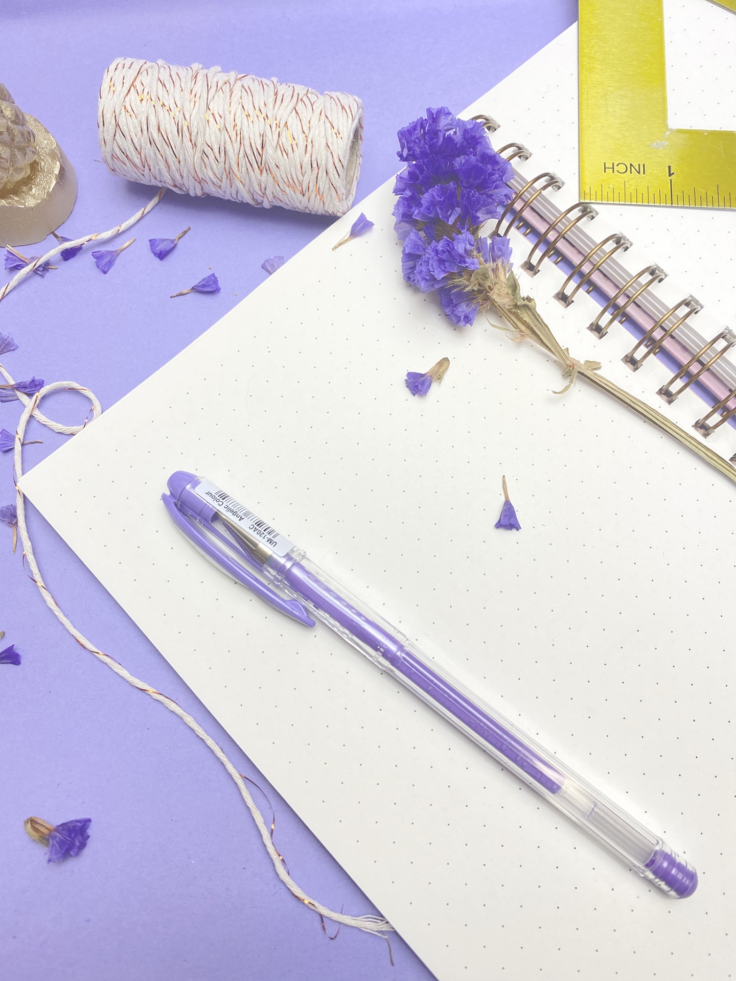 Caneta roxa transparente sobre caderno pontilhado com espiral e flores secas roxas