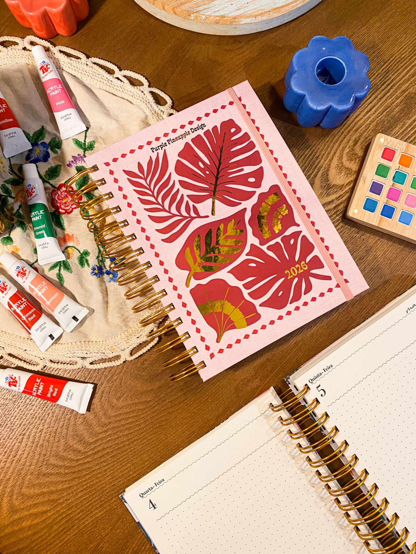 Caderno rosa com folhas decorativas, tubos de tinta acrílica, caderno aberto e castiçais cerâmicos