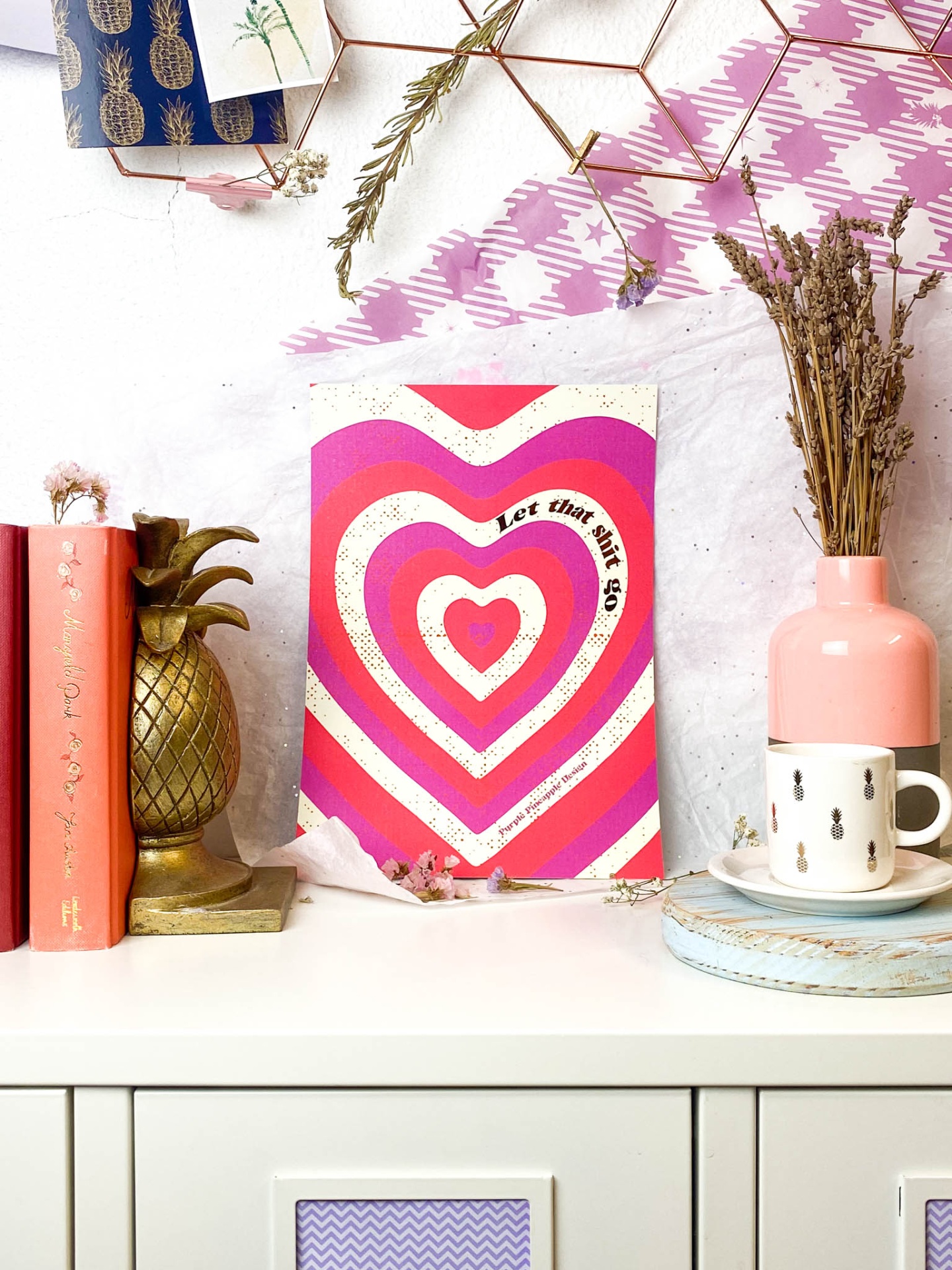 Superfície branca com livros cor de rosa, objeto decorativo dourado abacaxi, cartaz rosa e vermelho com texto, jarra e copo decorativos