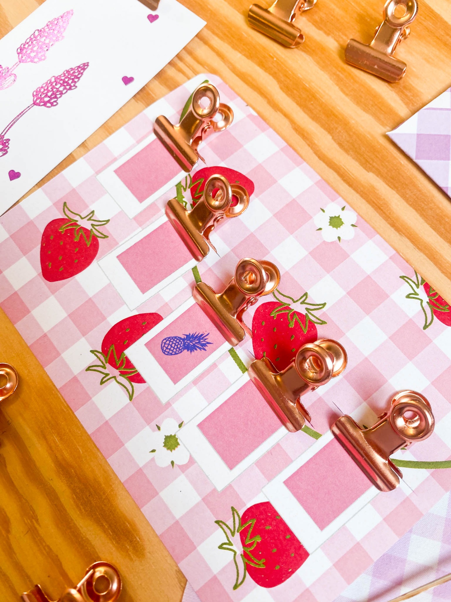 Pregadores metálicos dourados sobre papel rosa com ilustrações de morangos e flores em madeira clara