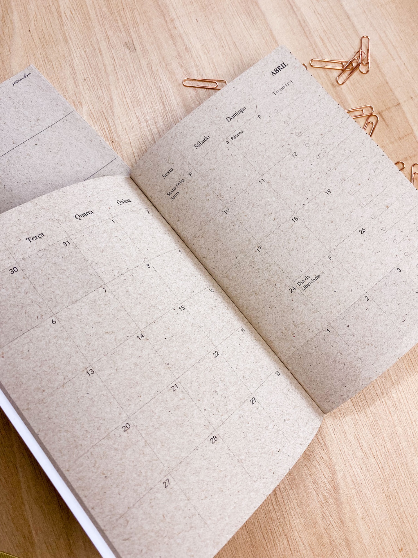 Caderno aberto com calendário em papel reciclado e clips de papel dourados sobre mesa de madeira