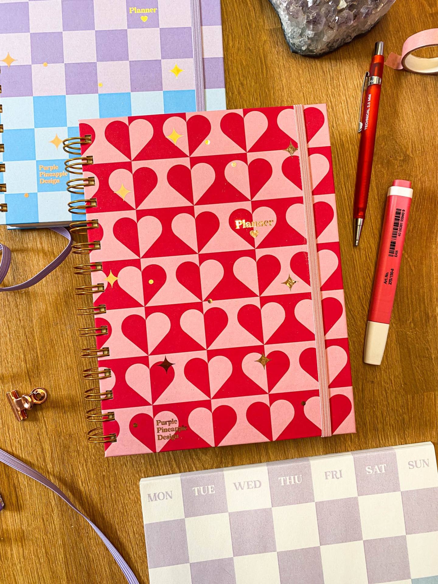 Caderno planner com padrão de corações rosa e vermelho, caneta e marcador ao lado sobre mesa de madeira