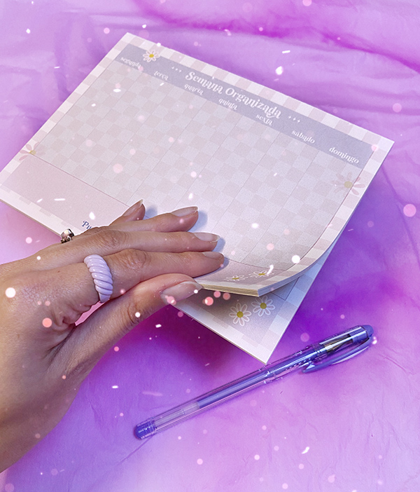 Bloco de notas semanal com design xadrez e flores, caneta e mão com anéis sobre superfície roxa