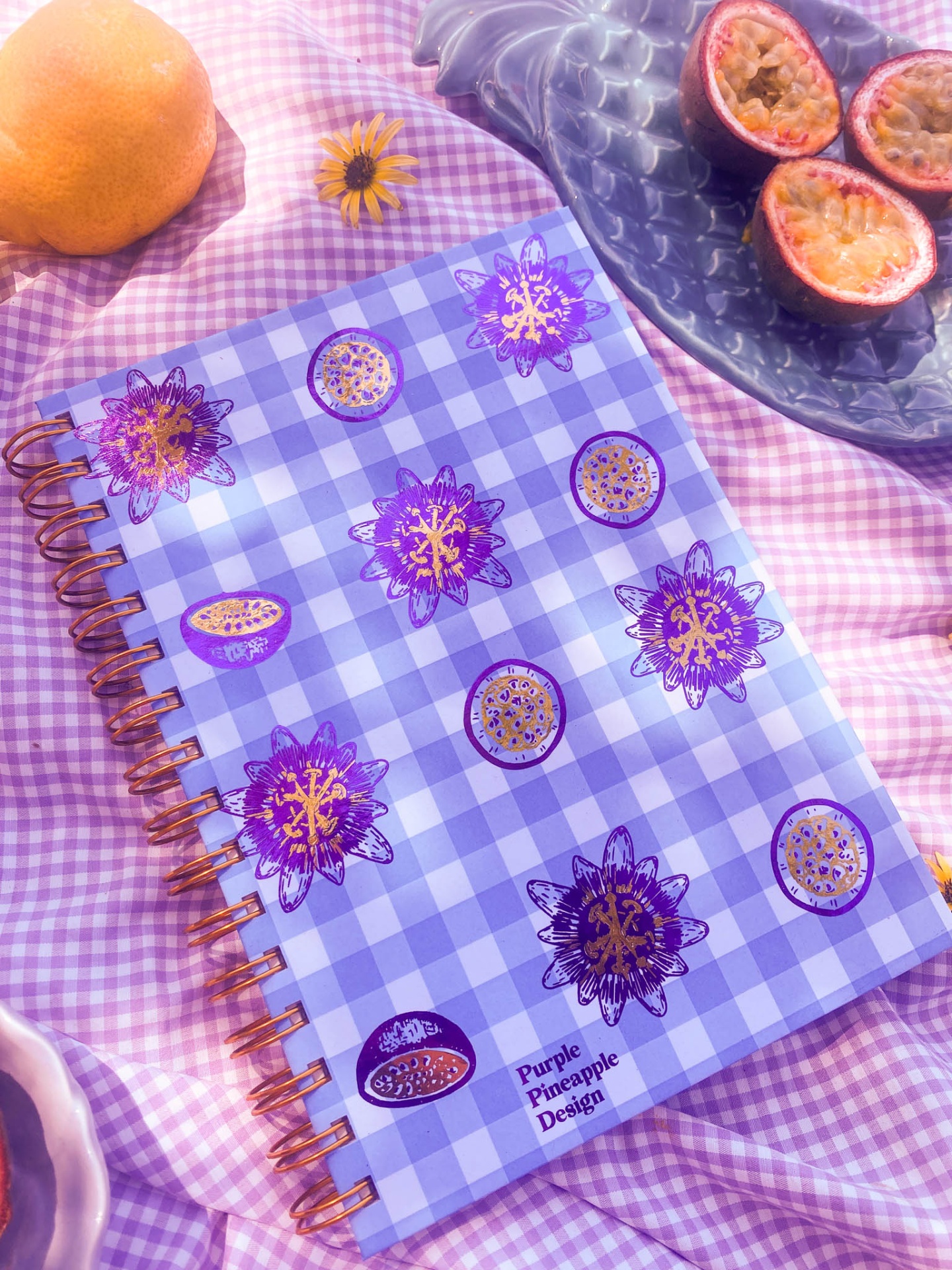 Caderno com capa xadrez azul e branco com desenhos de maracujá roxos e dourados, sobre tecido rosa, com frutas ao lado.