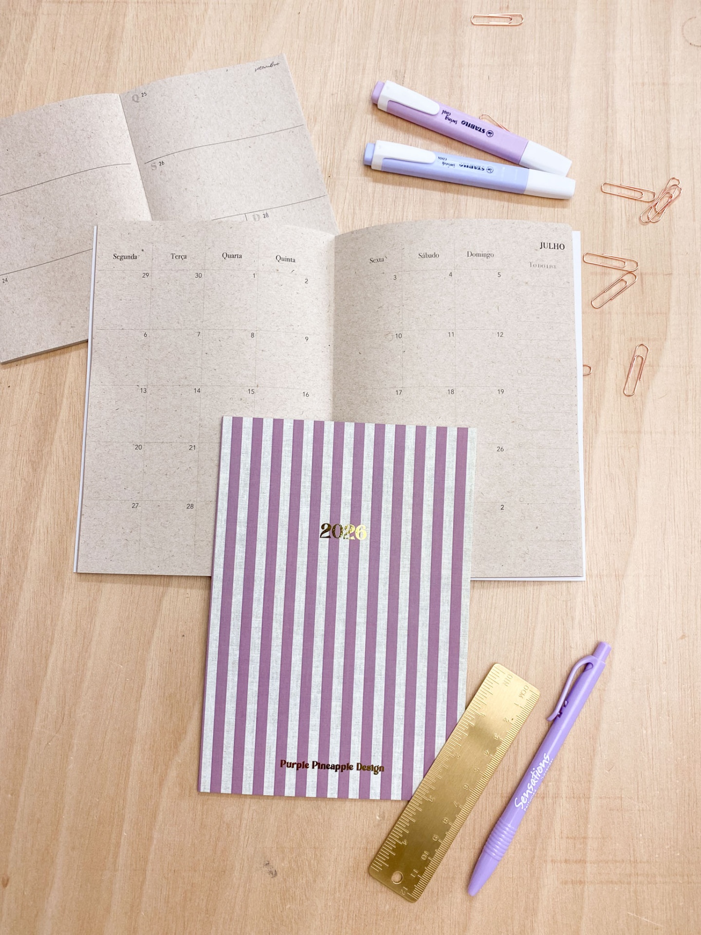 Agenda 2026 riscada lilás e branca com calendário, marcadores fluorescentes, caneta lilás e régua dourada sobre madeira
