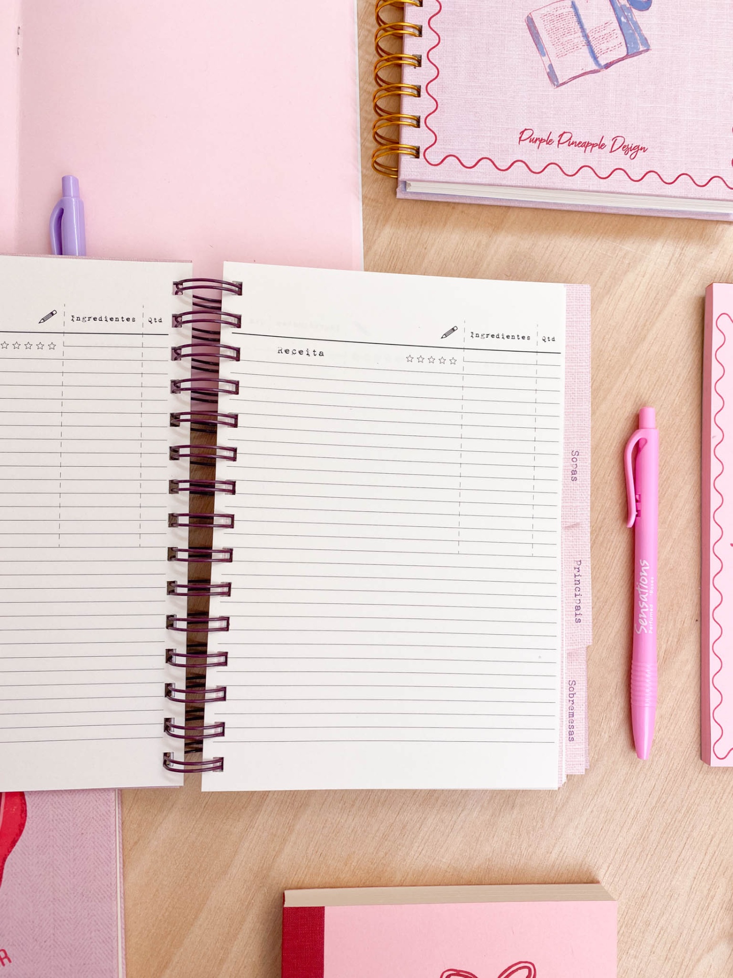 Caderno espiralado aberto para anotações com caneta rosa e outros cadernos sobre mesa de madeira