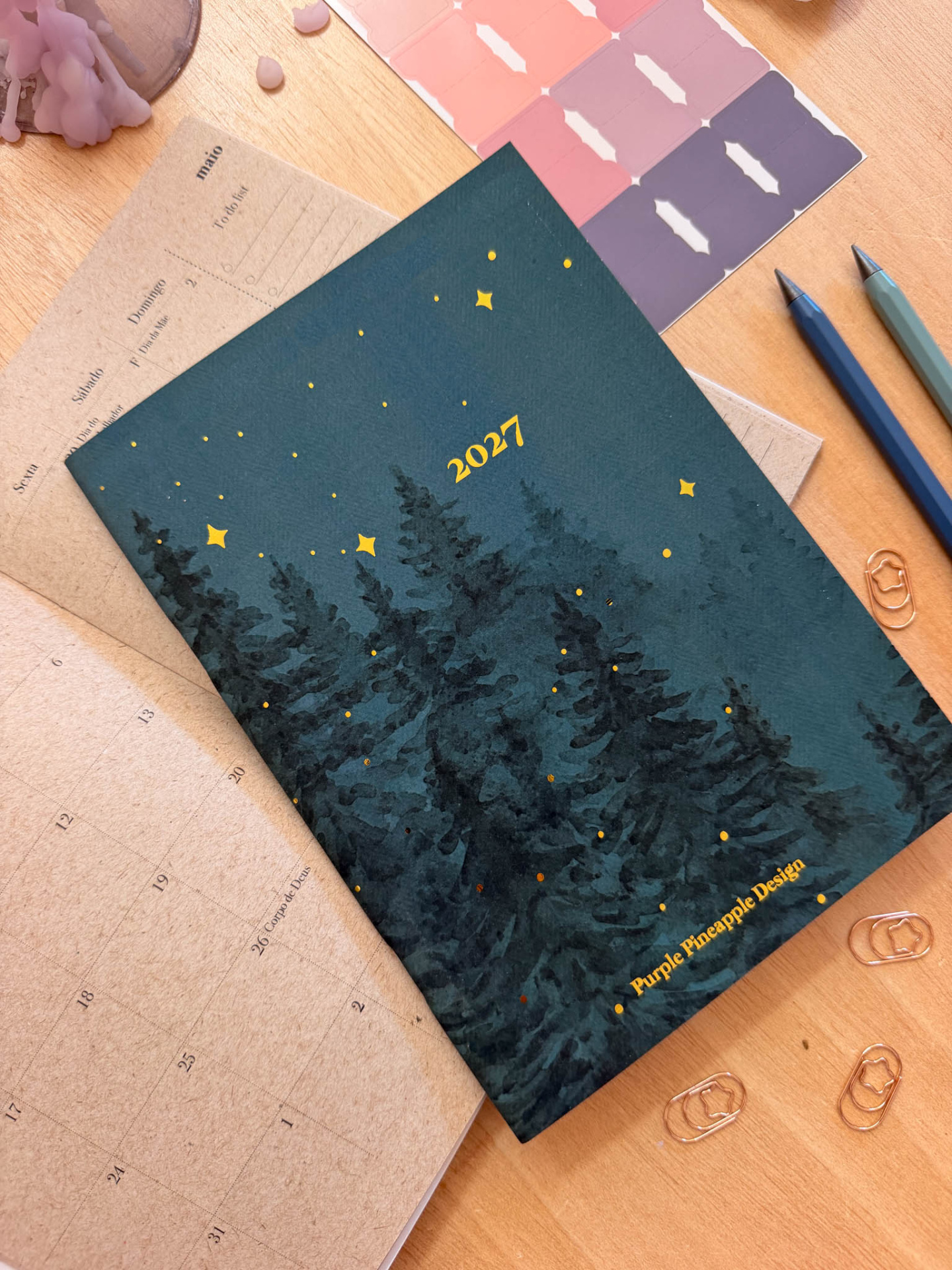 Calendário 2027 com capa azul e decoração de árvores e estrelas, sobre mesa de madeira com canetas e etiquetas adesivas