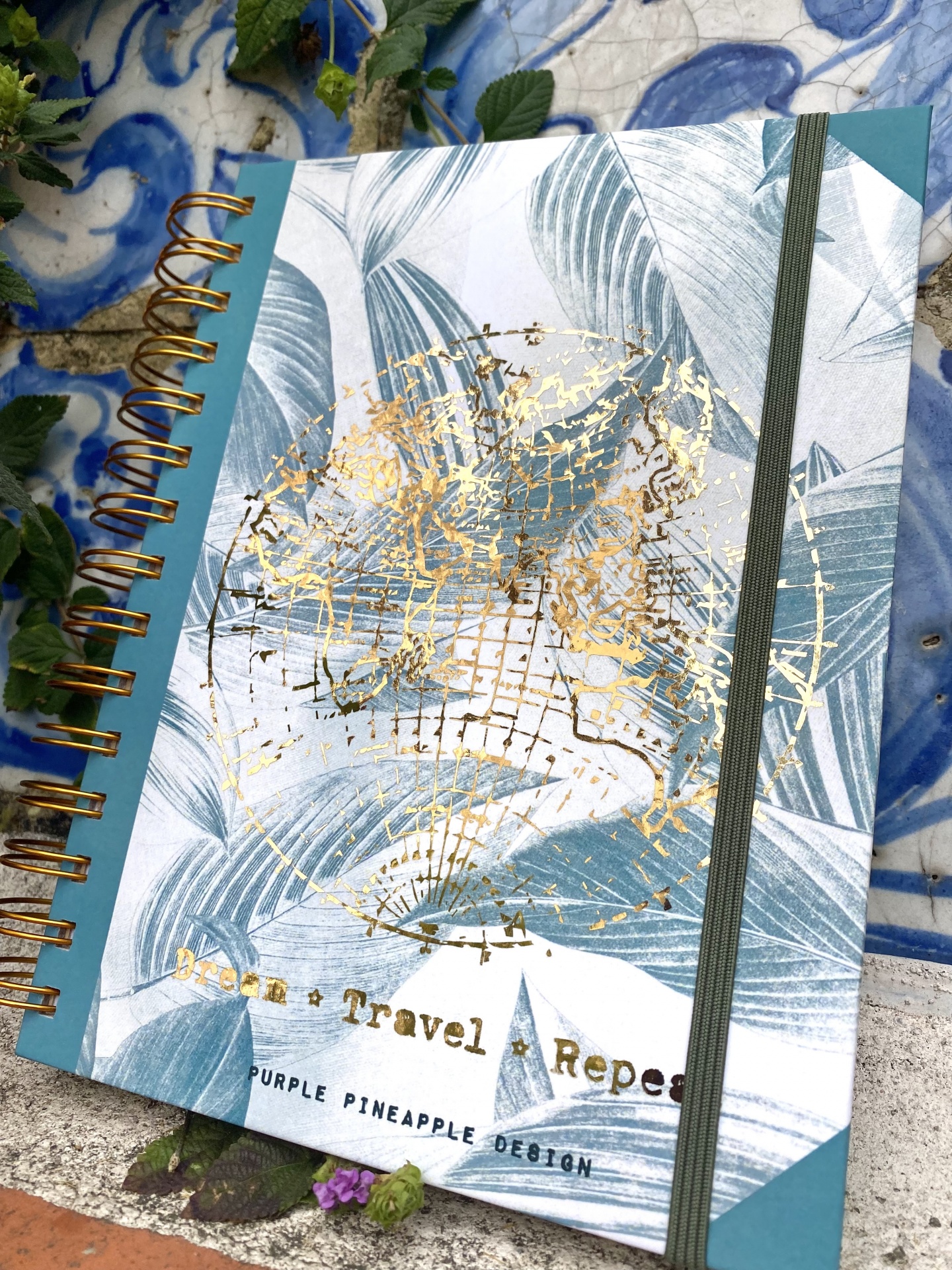 Caderno espiral com capa decorada de folhas azuladas e texto Travel Repeat