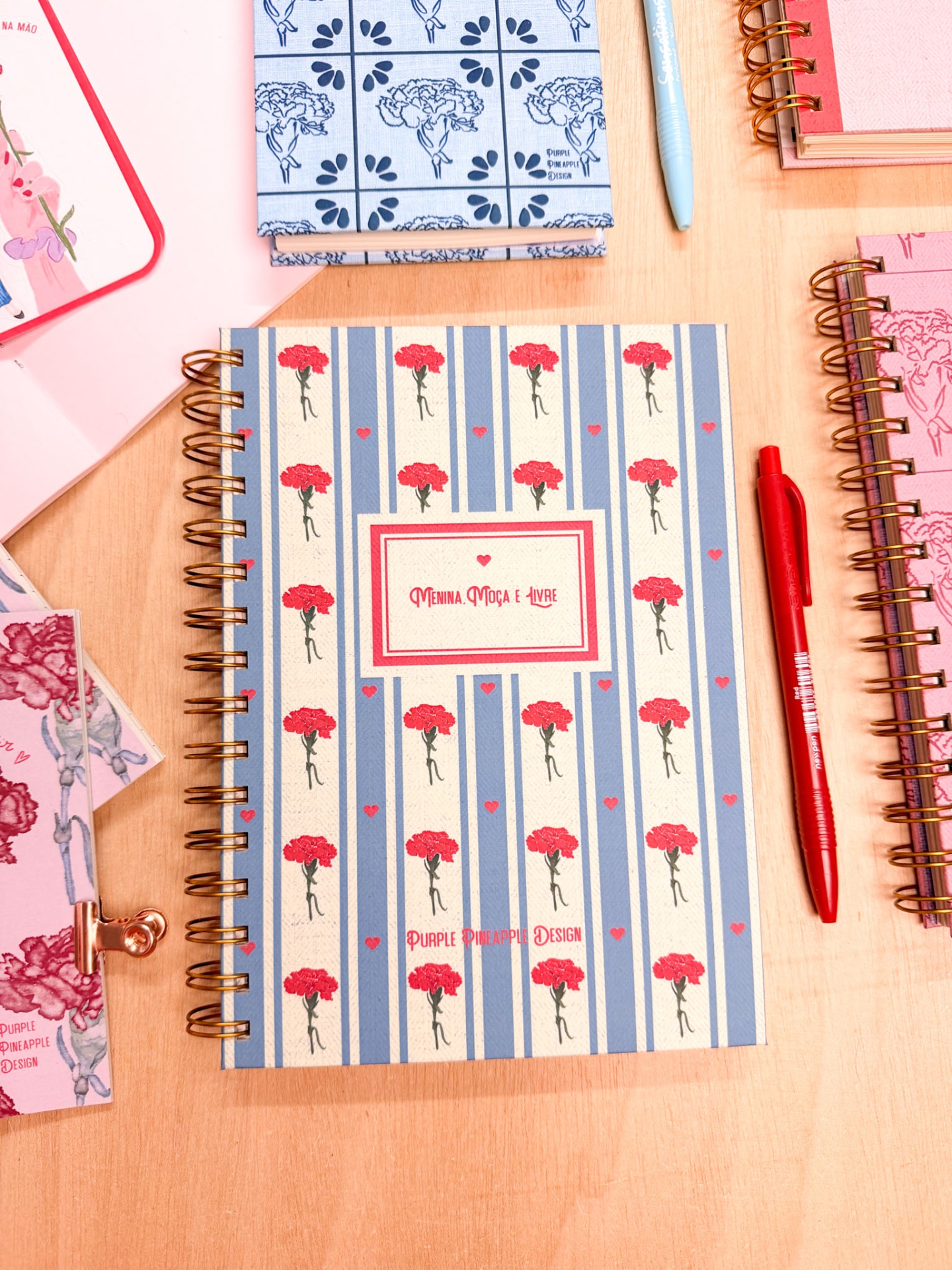 Caderno com capa azul e branca, flores vermelhas, caneta vermelha, sobre mesa de madeira