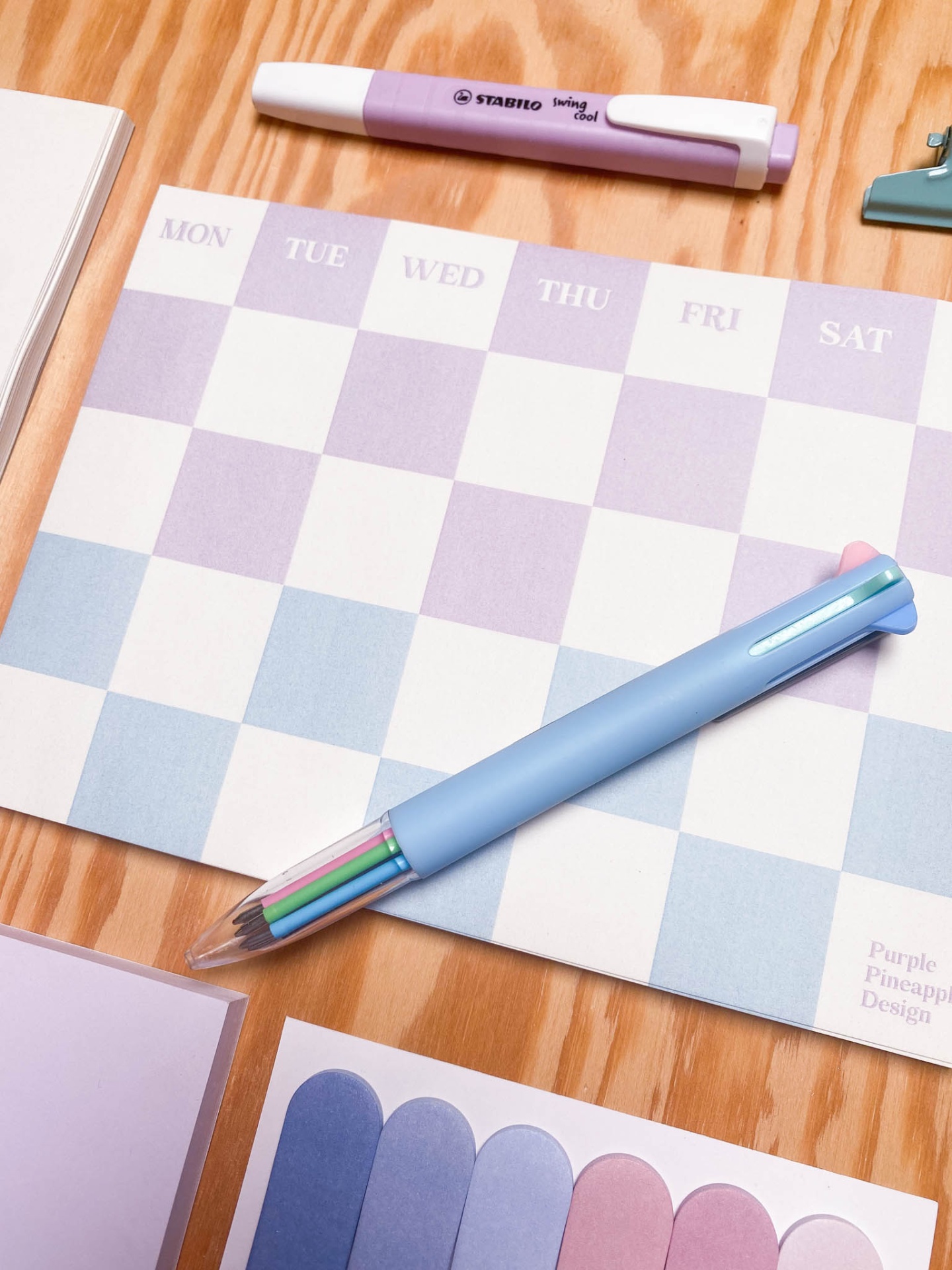 Agenda semanal lilás e azul com canetas e etiquetas coloridas sobre mesa de madeira