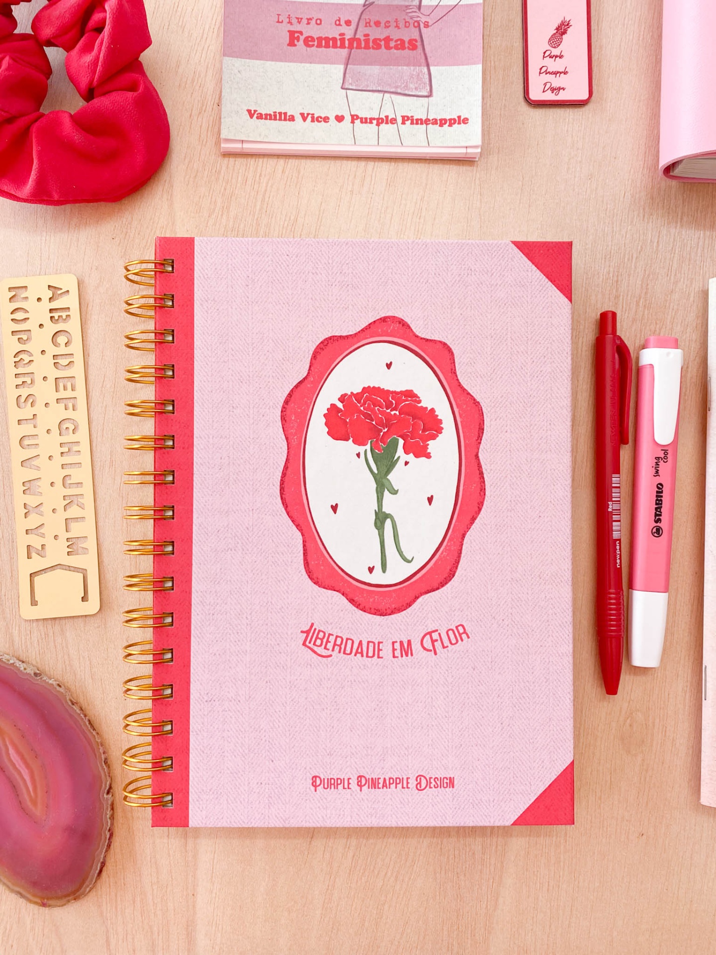 Caderno rosa e vermelho com flor, canetas e outros artigos de papelaria sobre mesa de madeira.