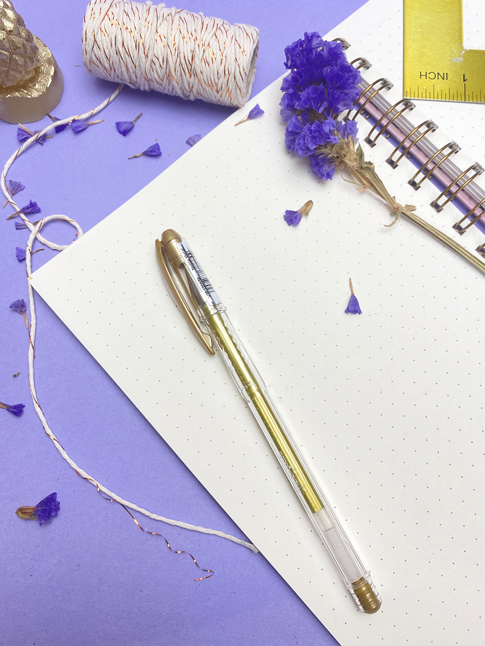 Caneta dourada sobre caderno espiralado pontilhado com flores lilases e régua amarela